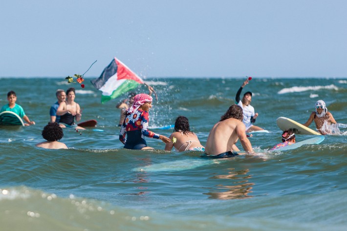 Surfers swim or sit on their boards in blue ocean water off the coast of Rockaway Beach 65 St. Someone holds a Palestinian flag while others throw flowers in the air during a paddle out.