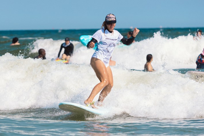 A surfer rides a small wave at Rockaway Beach while wearing a cap and a white shirt that reads "Gaza is a Beach Town"