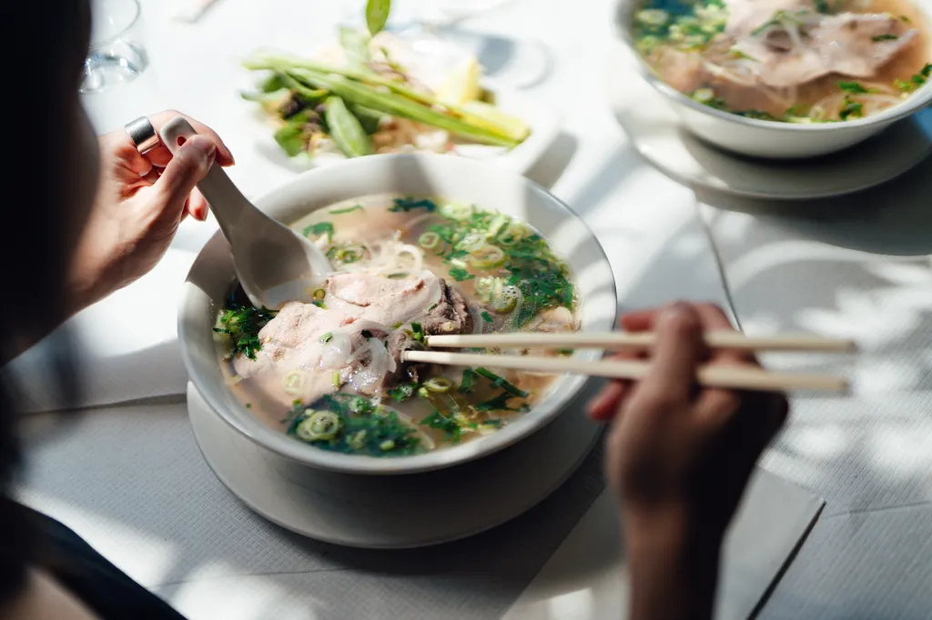 Two hands, one holding chopsticks and the other a wide spoon, hover over a bowl of pho.