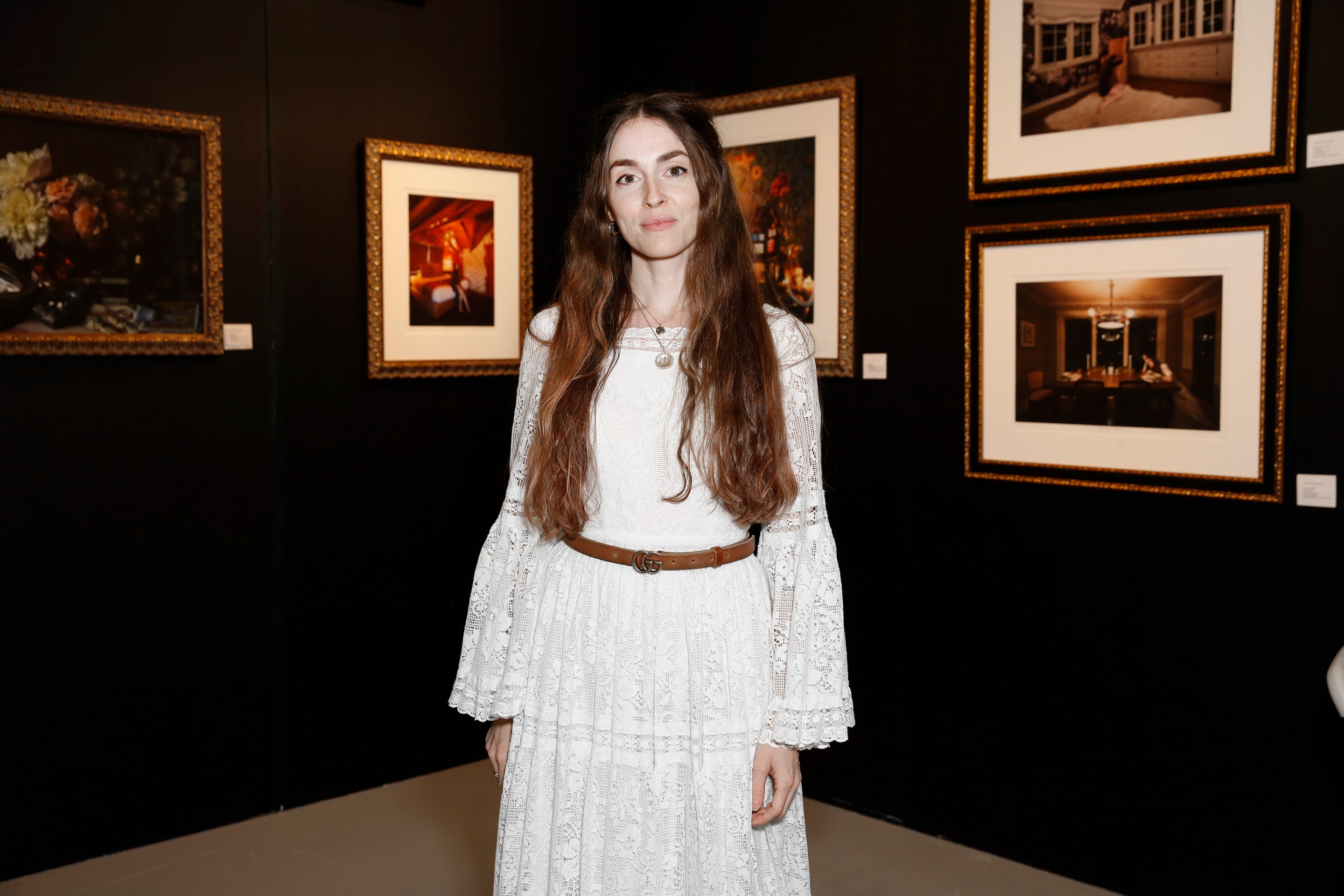 A woman with long brown hair stands in an art gallery, wearing a white lace dress with a brown belt. Behind her are framed photographs on dark walls, featuring various interior scenes. She smiles, looking at the camera.