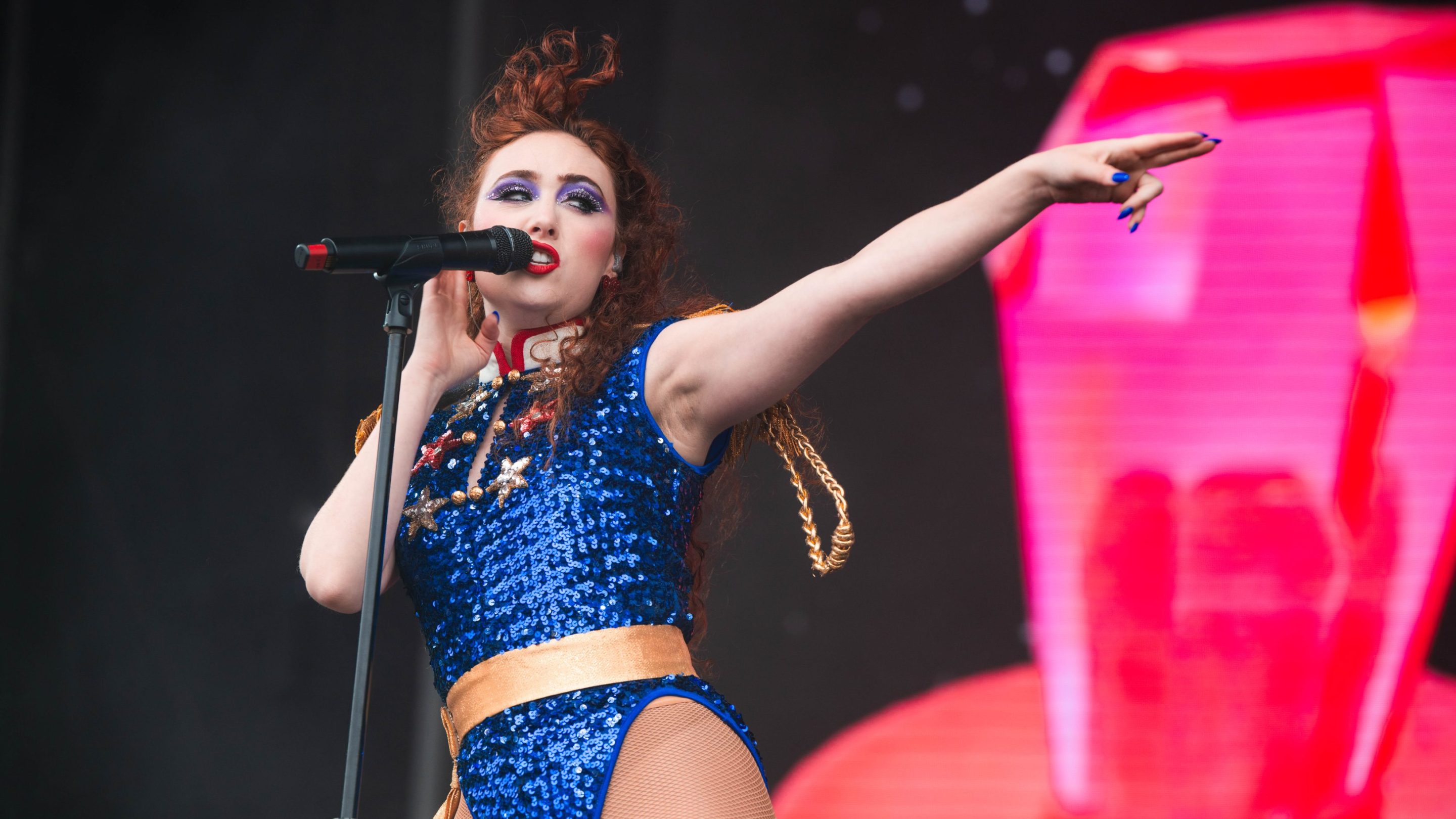 Chappell Roan on stage in a blue outfit, her hair blowing behind her, with one arm outstretched, pointing.