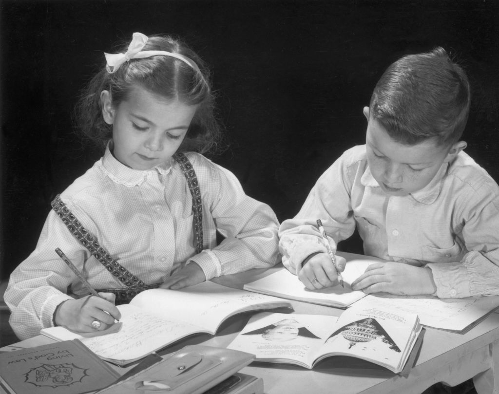 circa 1955: Two elementary school students complete homework assignments together, the girl has a book, 'Living By God's Law,' next to her on the table.