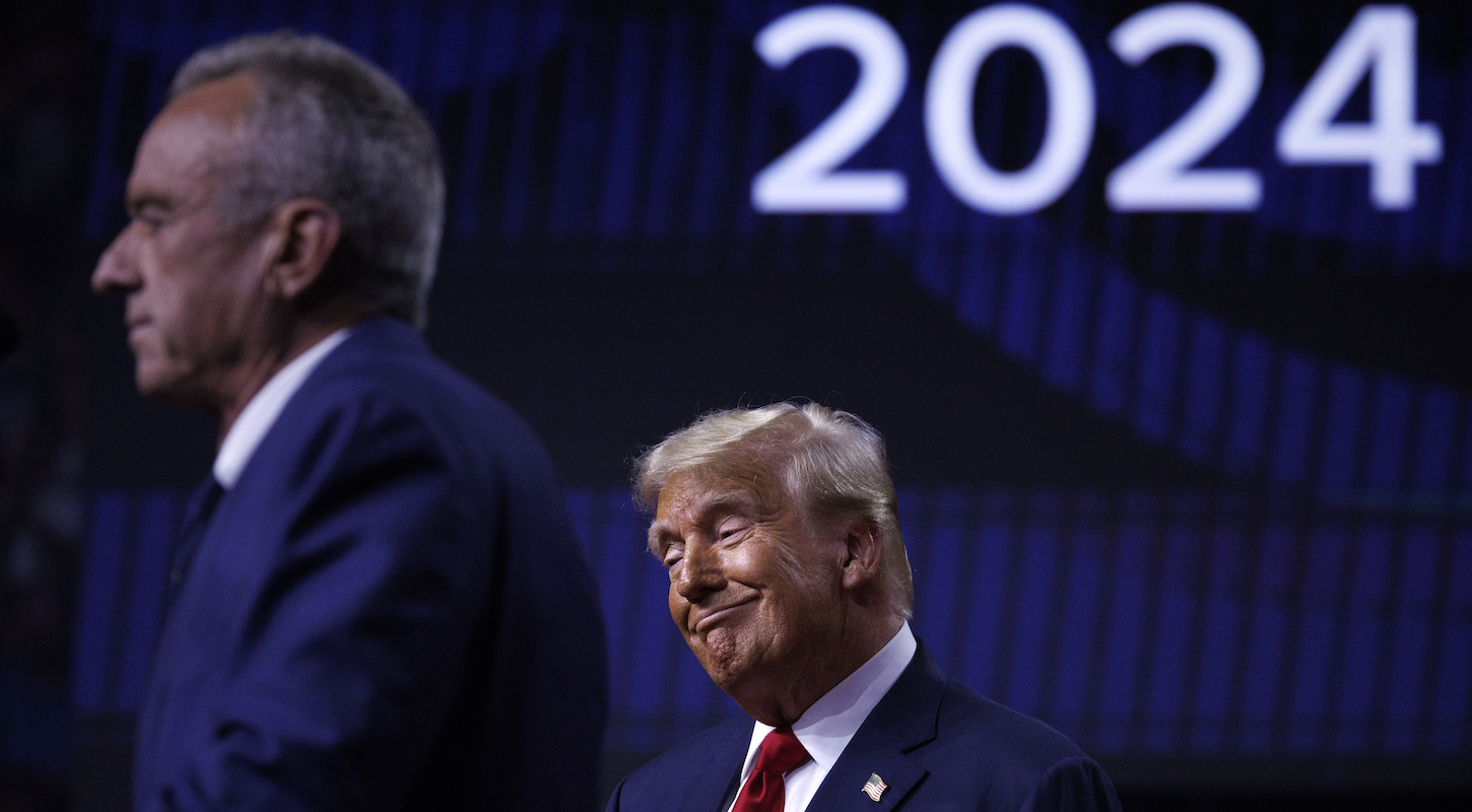 Donald Trump wears an ain't-I-a-stinker smirk while Robert F. Kennedy Jr. speaks onstage at a campaign event at which the latter announced the end of his presidential campaign and endorsement of the former.