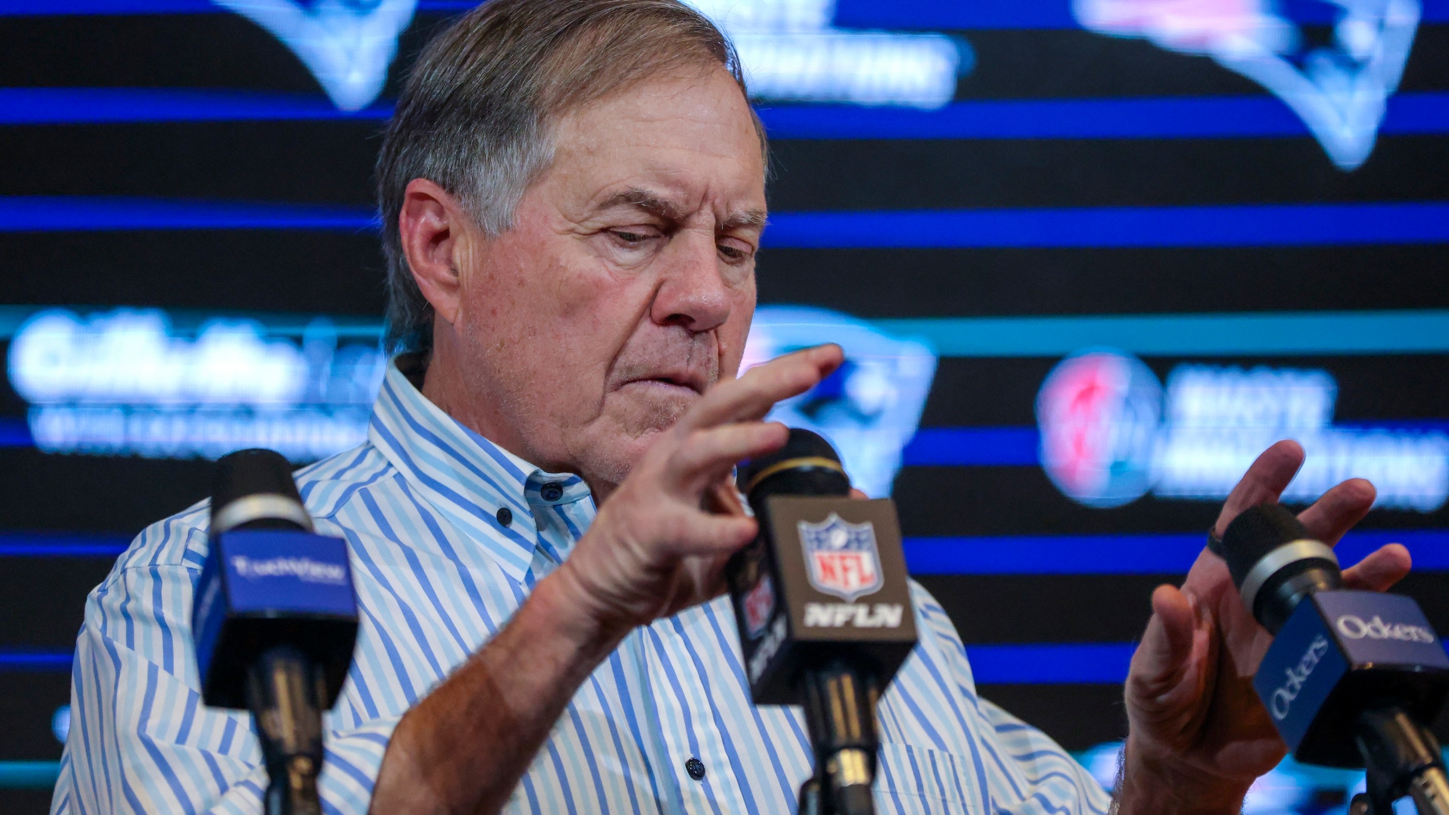 New England Patriots head coach Bill Belichick adjusts the microphones at his post-game press conference after the Patriots beat the Jets in January of 2024. He looks like he has a tummyache.