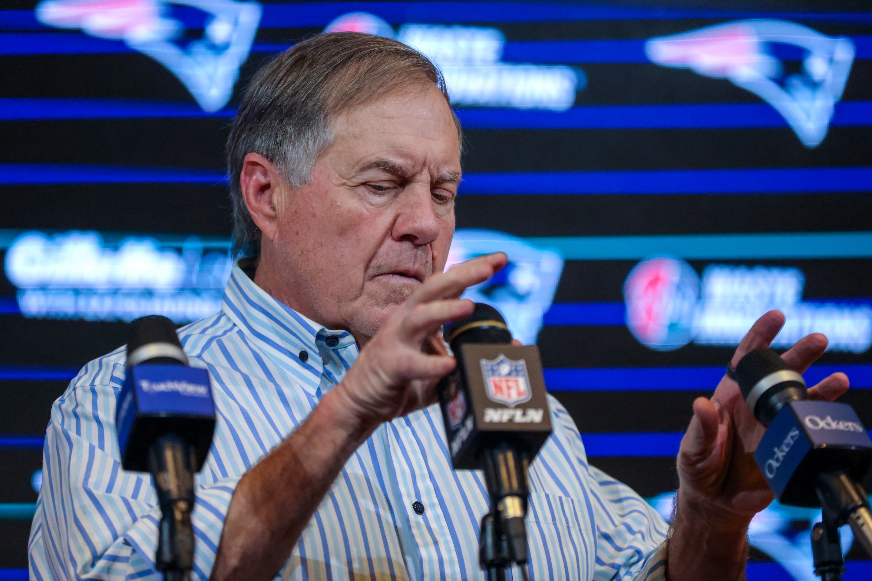 New England Patriots head coach Bill Belichick adjusts the microphones at his post-game press conference after the Patriots beat the Jets in January of 2024. He looks like he has a tummyache.