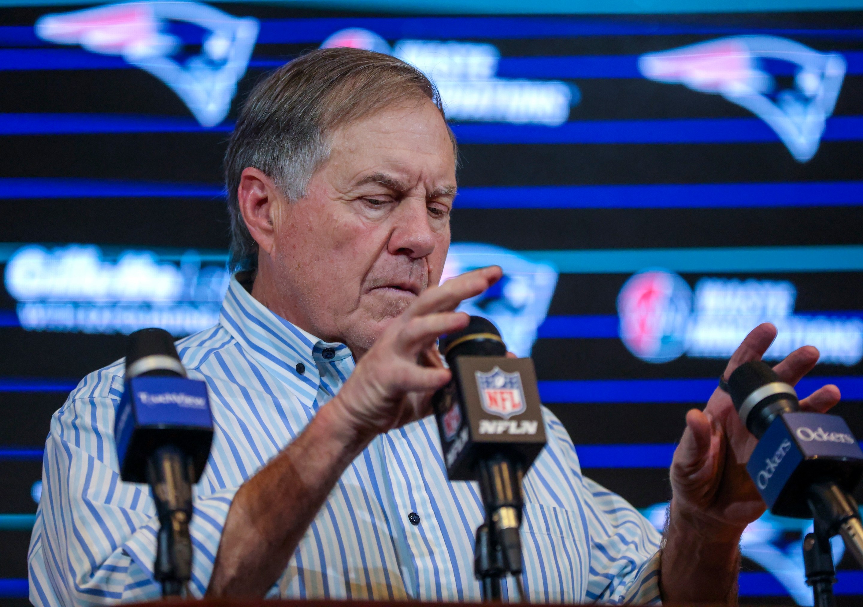 New England Patriots head coach Bill Belichick adjusts the microphones at his post-game press conference after the Patriots beat the Jets in January of 2024. He looks like he has a tummyache.