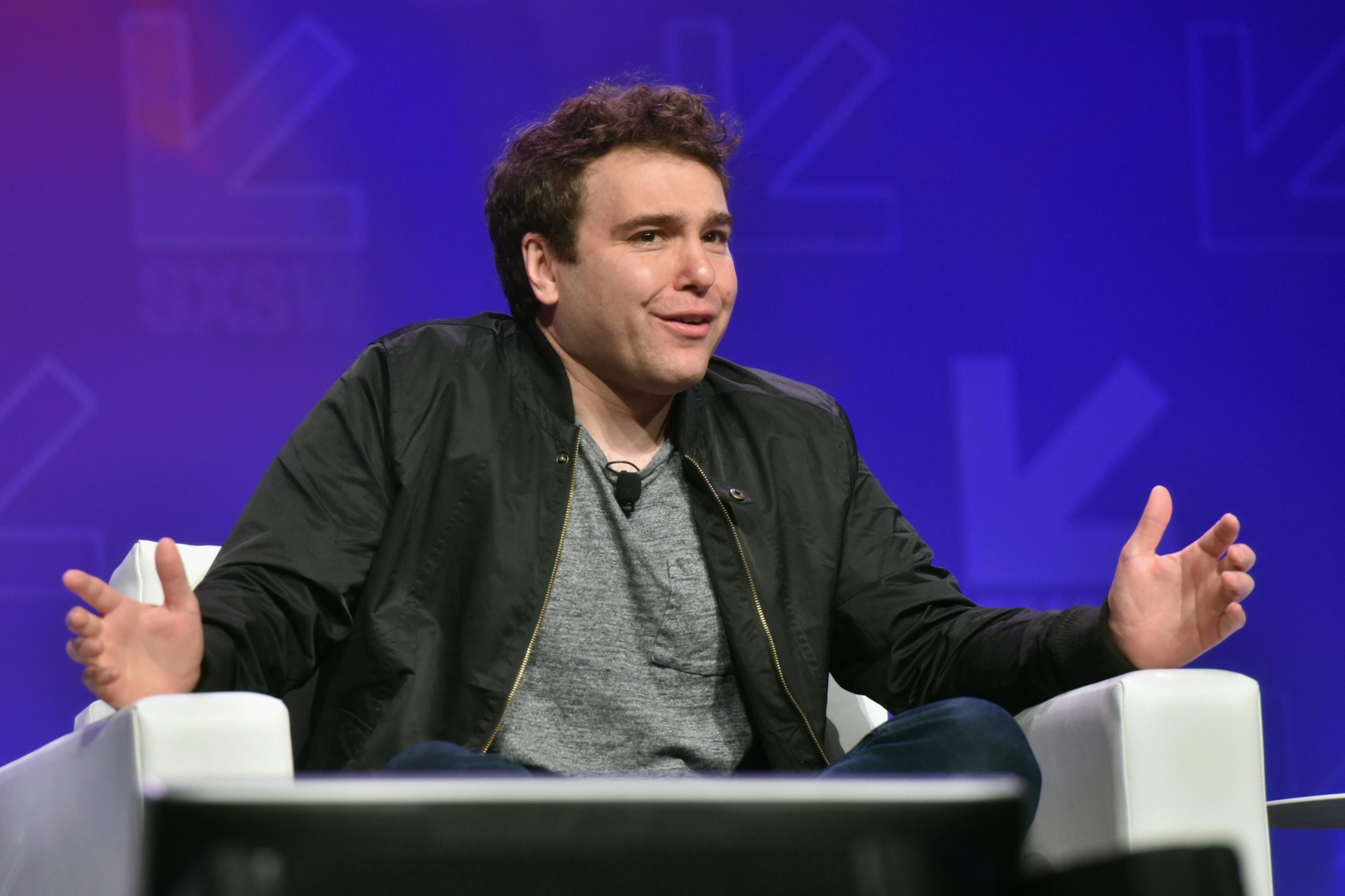 Jon Lovett sits on stage in a chair, cross-legged. He holds his hands apart and looks at the audience with a sort of chagrined expression.