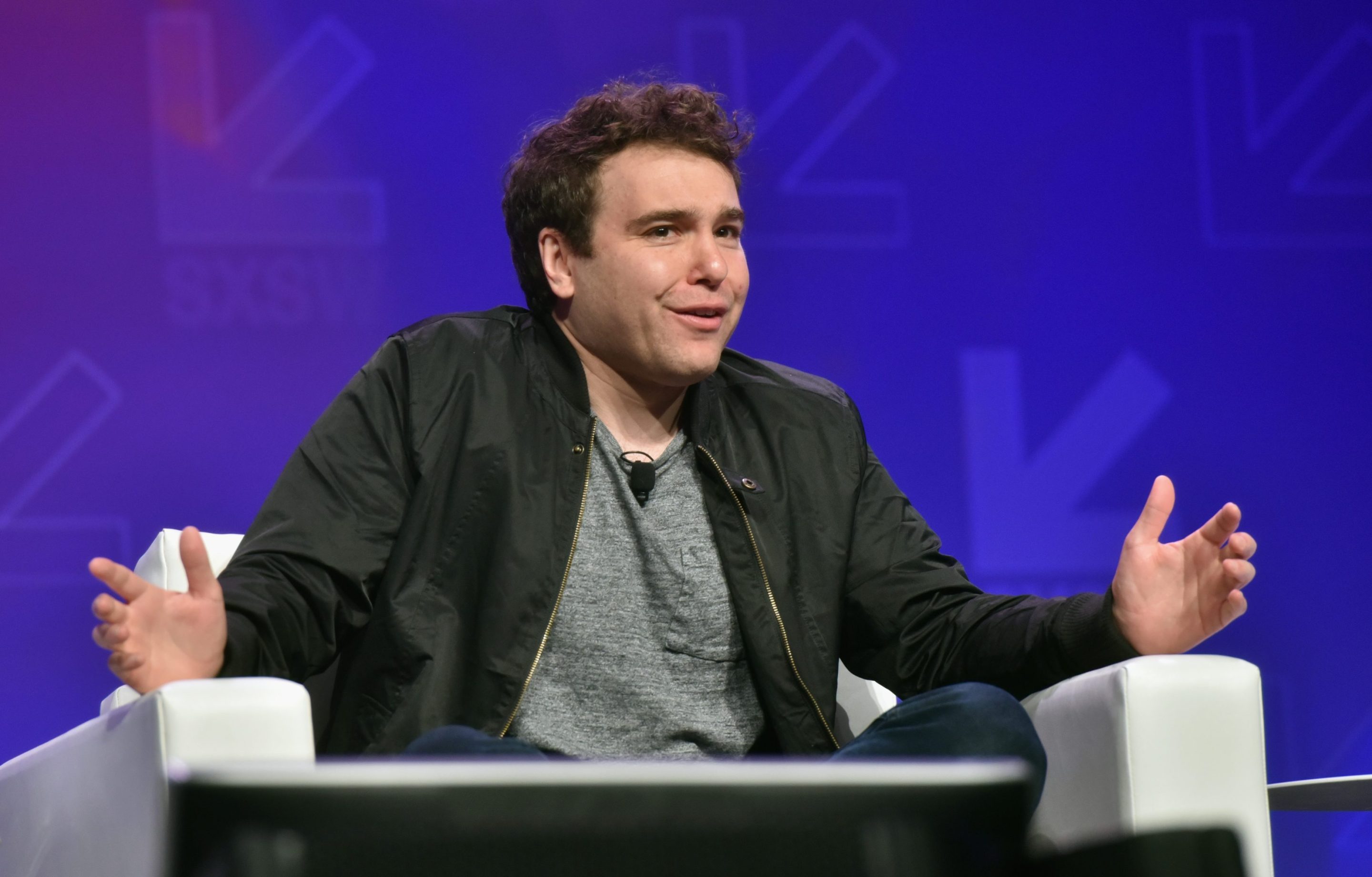 Jon Lovett sits on stage in a chair, cross-legged. He holds his hands apart and looks at the audience with a sort of chagrined expression.