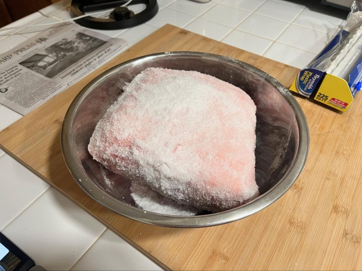 A big hunk of pork, coated in sugar and salt, in an aluminum mixing bowl.