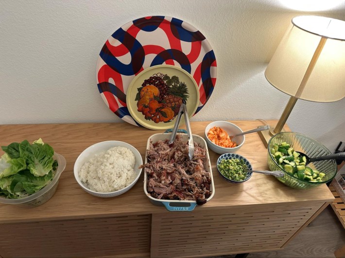 A sideboard-type piece of furniture with a spread of food including pork, rice, lettuce, cucumber salad, and kimchi, plus a pair of decorative plates and a lamp.