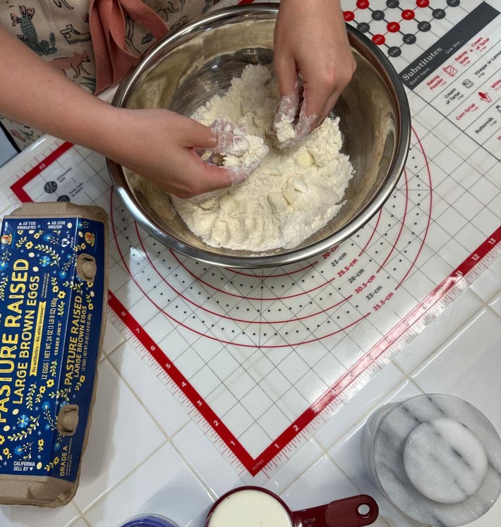 In a large aluminum mixing bowl, a pair of hands pinch cold butter into dry scone ingredients.