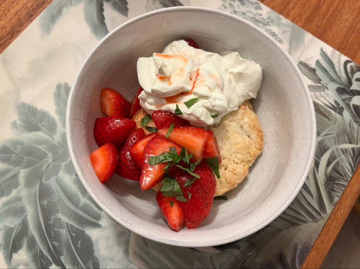 A white ceramic bowl containing a scone, some whipped mascarpone cream, and some macerated strawberries, with a scattering of chopped mint over the top.