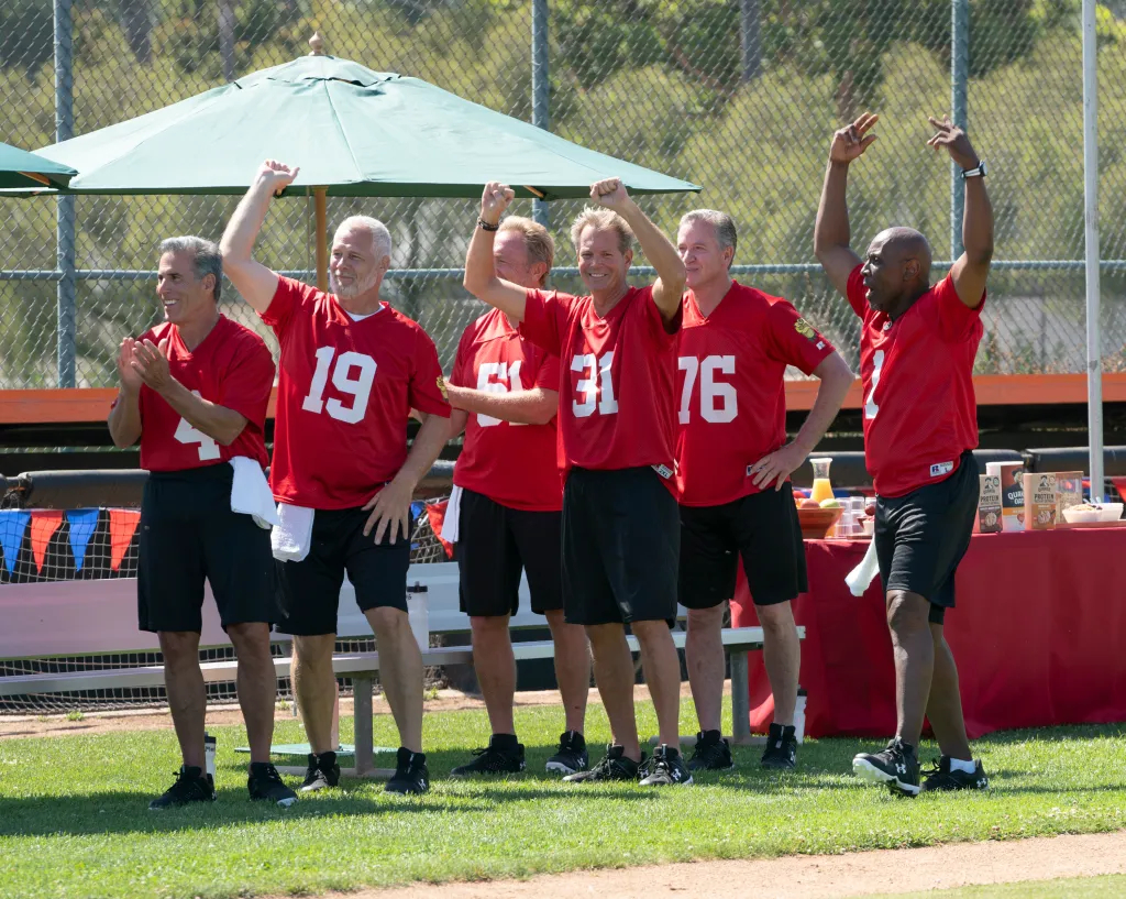 Six older gentlemen wearing red jerseys stand on a green field, cheering excitedly.