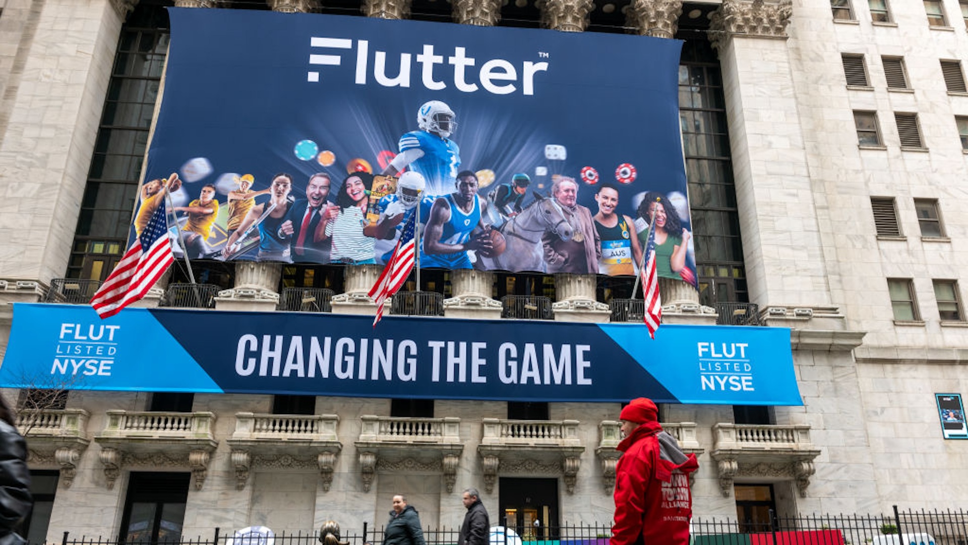 People walk by a banner outside of the New York Stock Exchange (NYSE) for the IPO of Flutter Entertainment, the parent company of FanDuel, on January 29, 2024 in New York City. The company, which is already on the London Stock Exchange, will be listed under the “FLUT” symbol. Wall Street observers are watching to see if an upcoming Fed announcement on rate policy and the most recent violence in the Middle East will have any effect on the markets.