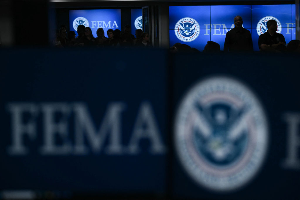 A look inside the offices of the Federal Emergency Management Agency, a sign for FEMA sits in the foreground as we see the silhouettes of people against large monitors in the background.