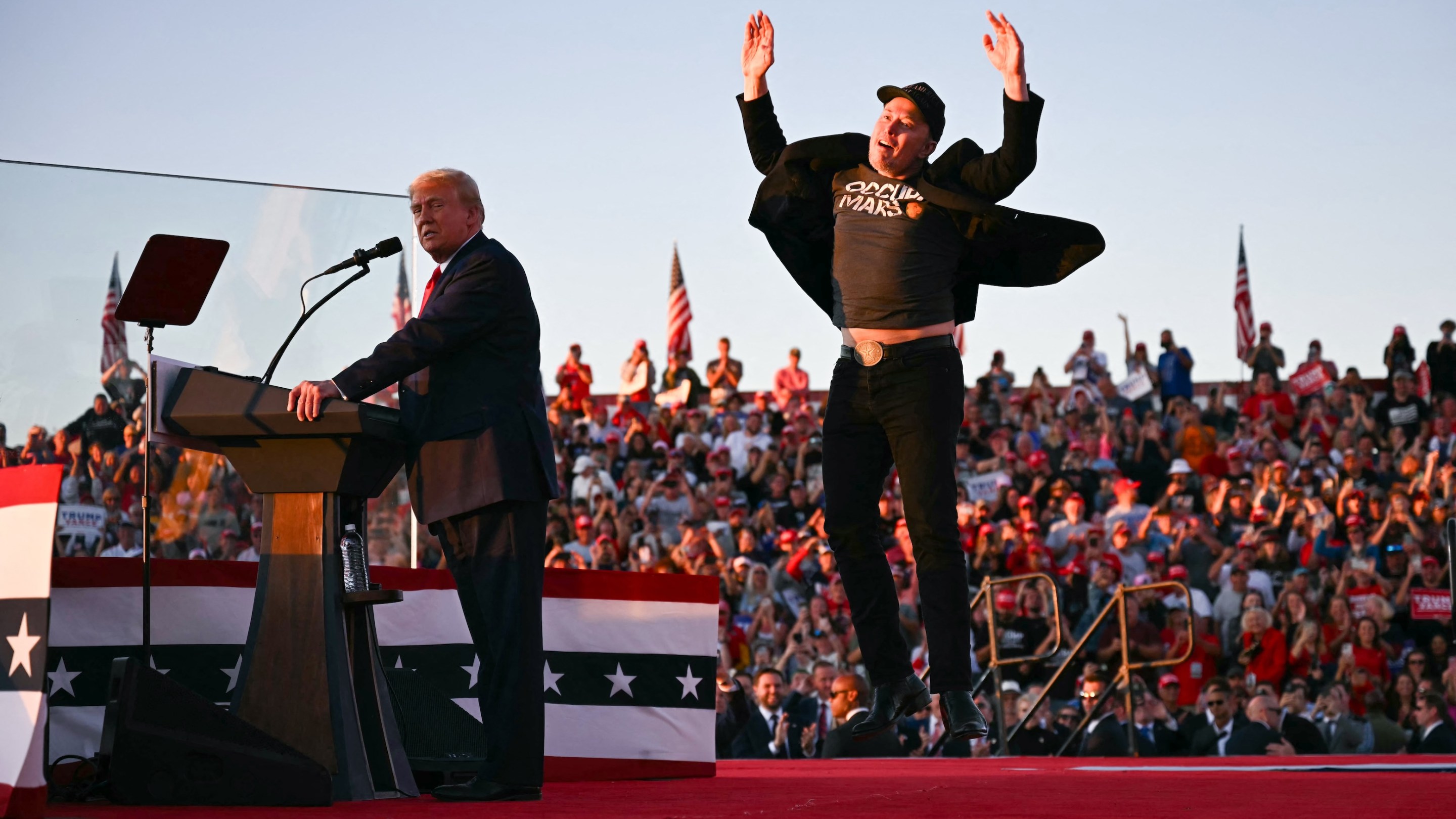 Tesla CEO Elon Musk (R) jumps on stage as he joins Donald Trump. You can see Musk's tummy; Trump looks kind of upset.