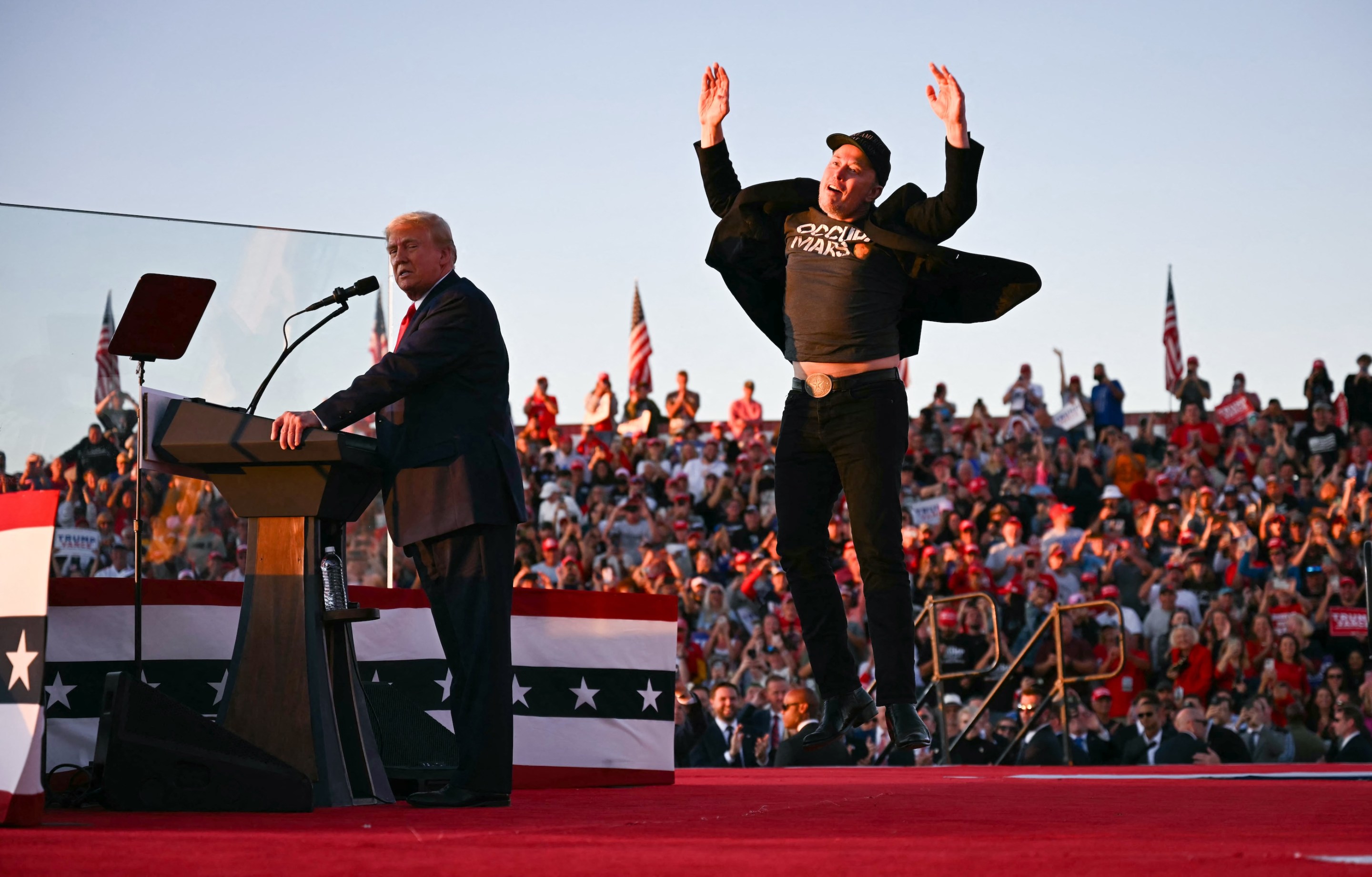 Tesla CEO Elon Musk (R) jumps on stage as he joins Donald Trump. You can see Musk's tummy; Trump looks kind of upset.