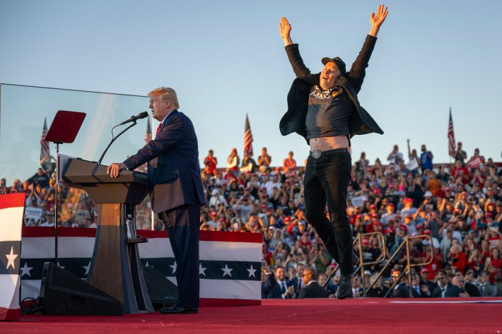 Donald Trump standing behind a podium at his rally in Butler, Pennsylvania while Elon Musk jumps around weirdly behind him. In this case his arms are extended over his head and you can see his tummy.