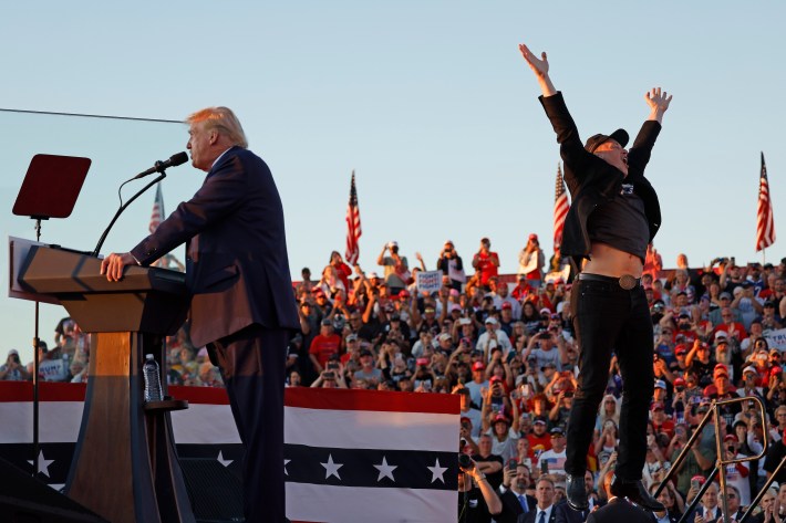 Elon Musk leaps on stage with Donald Trump in Butler, Pennsylvania. Trump is talking and Musk is jumping in the air with his mouth open.