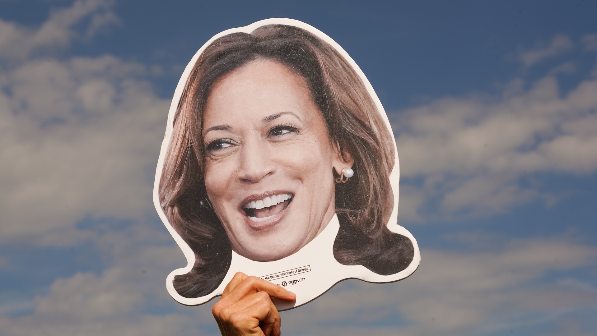 A big campaign sign that is just Kamala Harris's head, in front of a blue sky with scattered clouds.