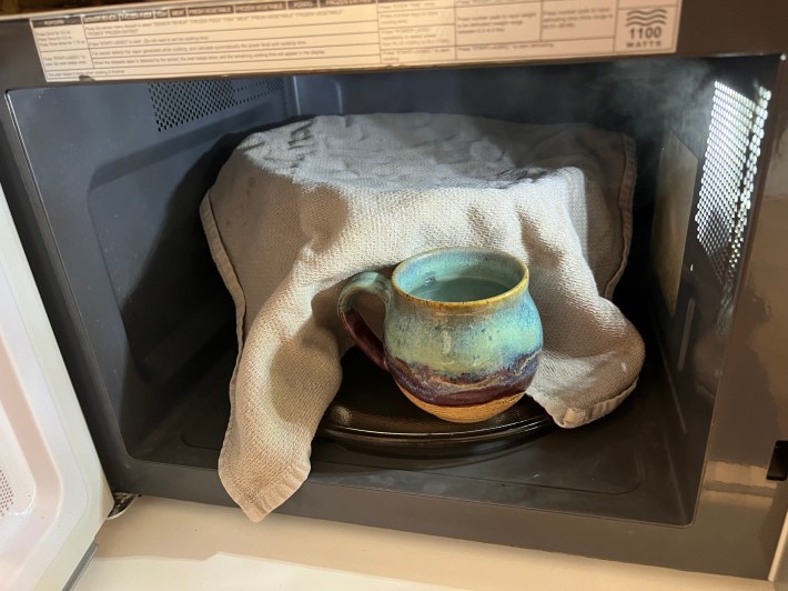 A large mixing bowl covered in a rag, inside a microwave, beside a mug of steaming water.