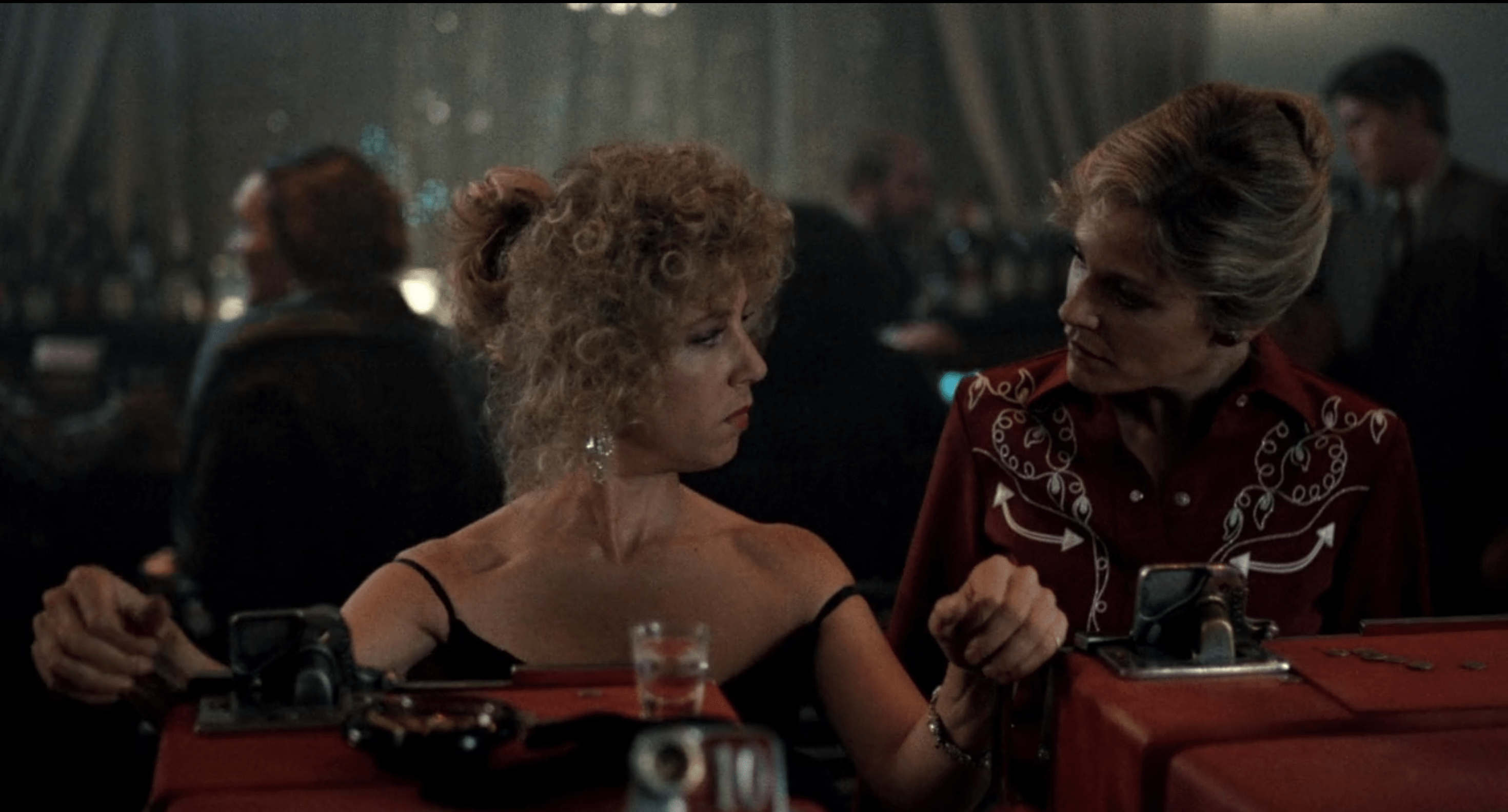 A screenshot of two blonde women looking at each other, one playing the slots, in a 60s-era casino