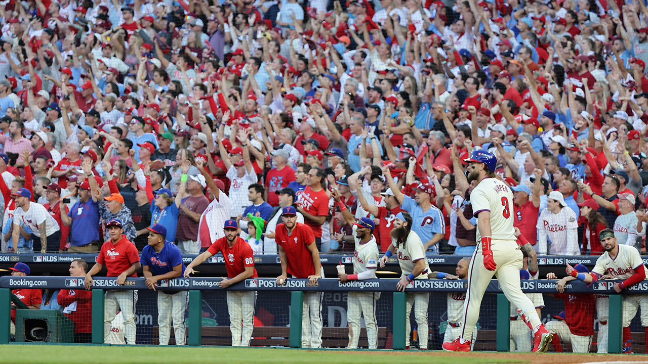 Long angle shot of Bryce Harper standing there, back 3/4 to the camera, looking at the ball. He's just hit a homer. Behind him in the photo is the Phillies dugout watching, and fans going wild in the stands. It's a long shot, about 25-30 rows of fans.