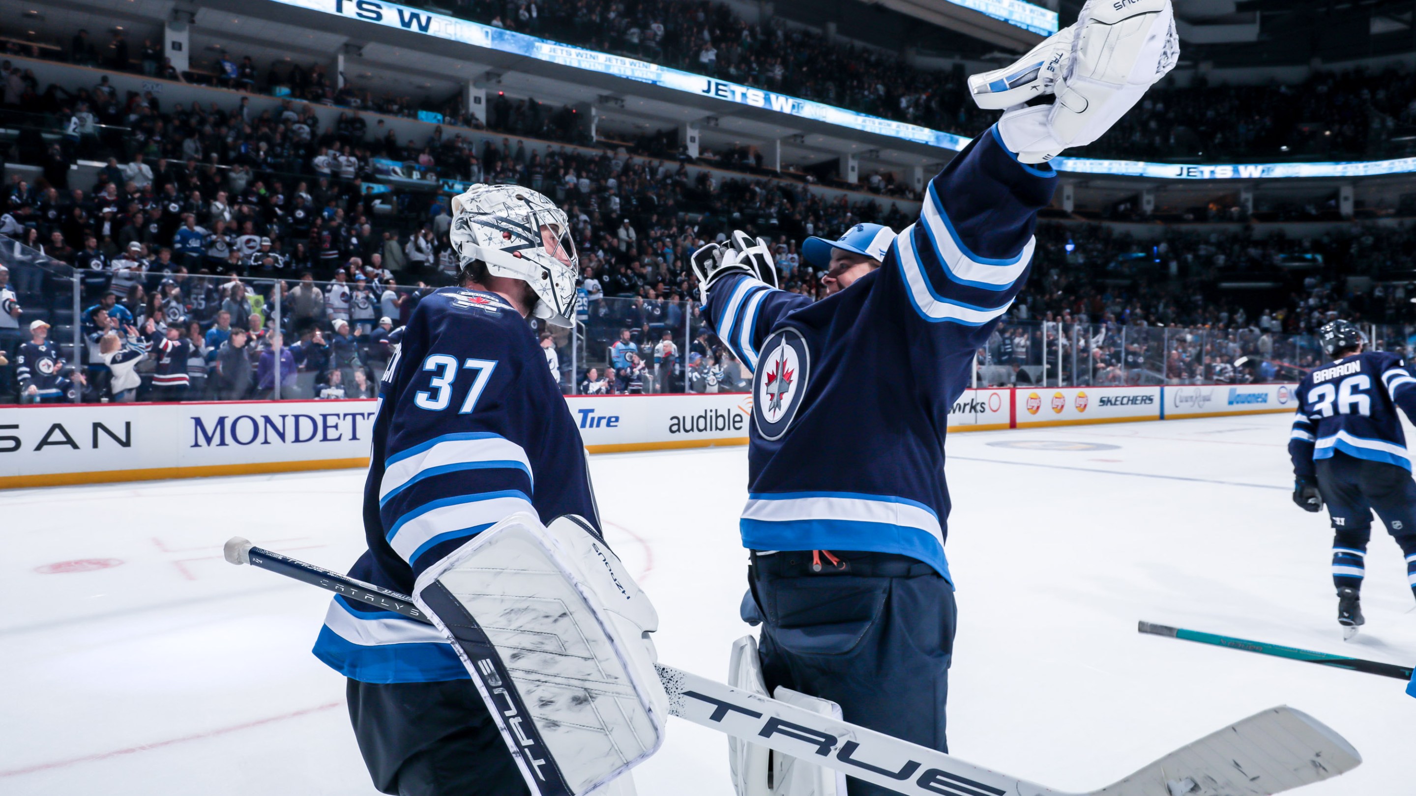 Goaltenders Connor Hellebuyck (left, in a helmet) and Eric Comrie (right, arms flung up as if to give a big hug) of the Winnipeg Jets celebrate following a 7-4 victory over the Tampa Bay Lightning.