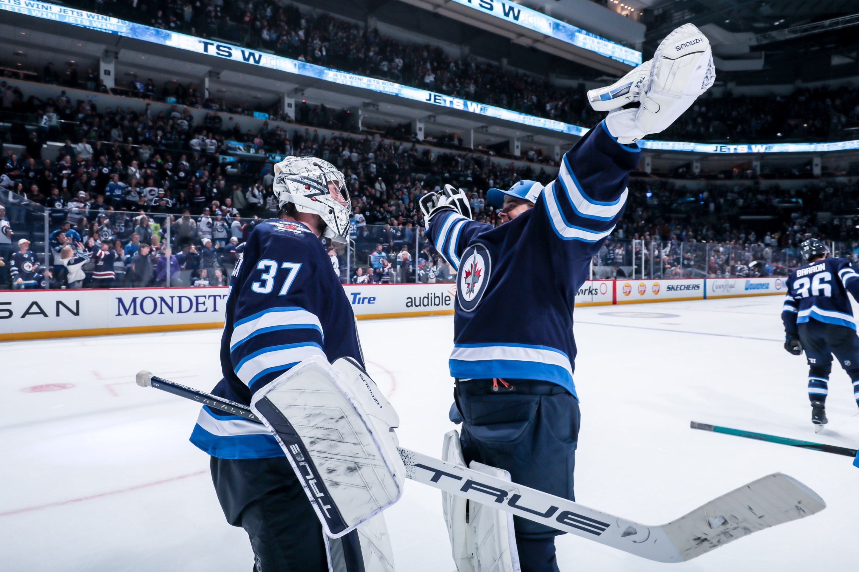 Goaltenders Connor Hellebuyck (left, in a helmet) and Eric Comrie (right, arms flung up as if to give a big hug) of the Winnipeg Jets celebrate following a 7-4 victory over the Tampa Bay Lightning.