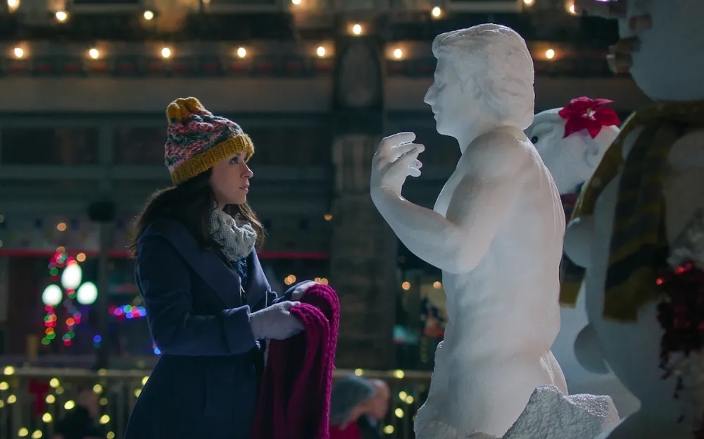 a still for the netflix movie hot frosty: lacey chabert in a beanie stands in front of a sexy snowman, holding a scarf