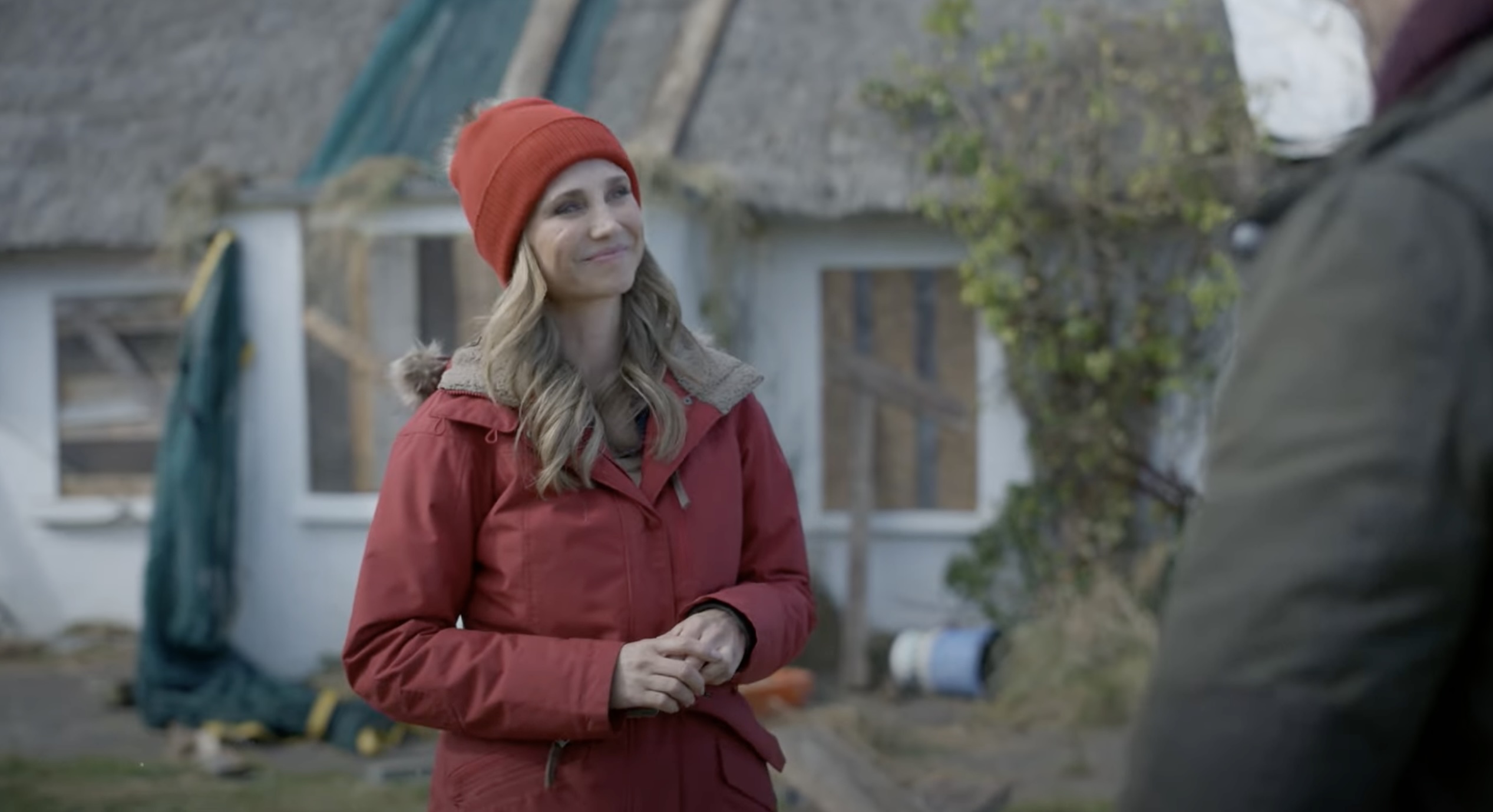 A still from a movie. a white woman in a red coat and a red beanie stands in front of a dilapidated house that needs fixing up.