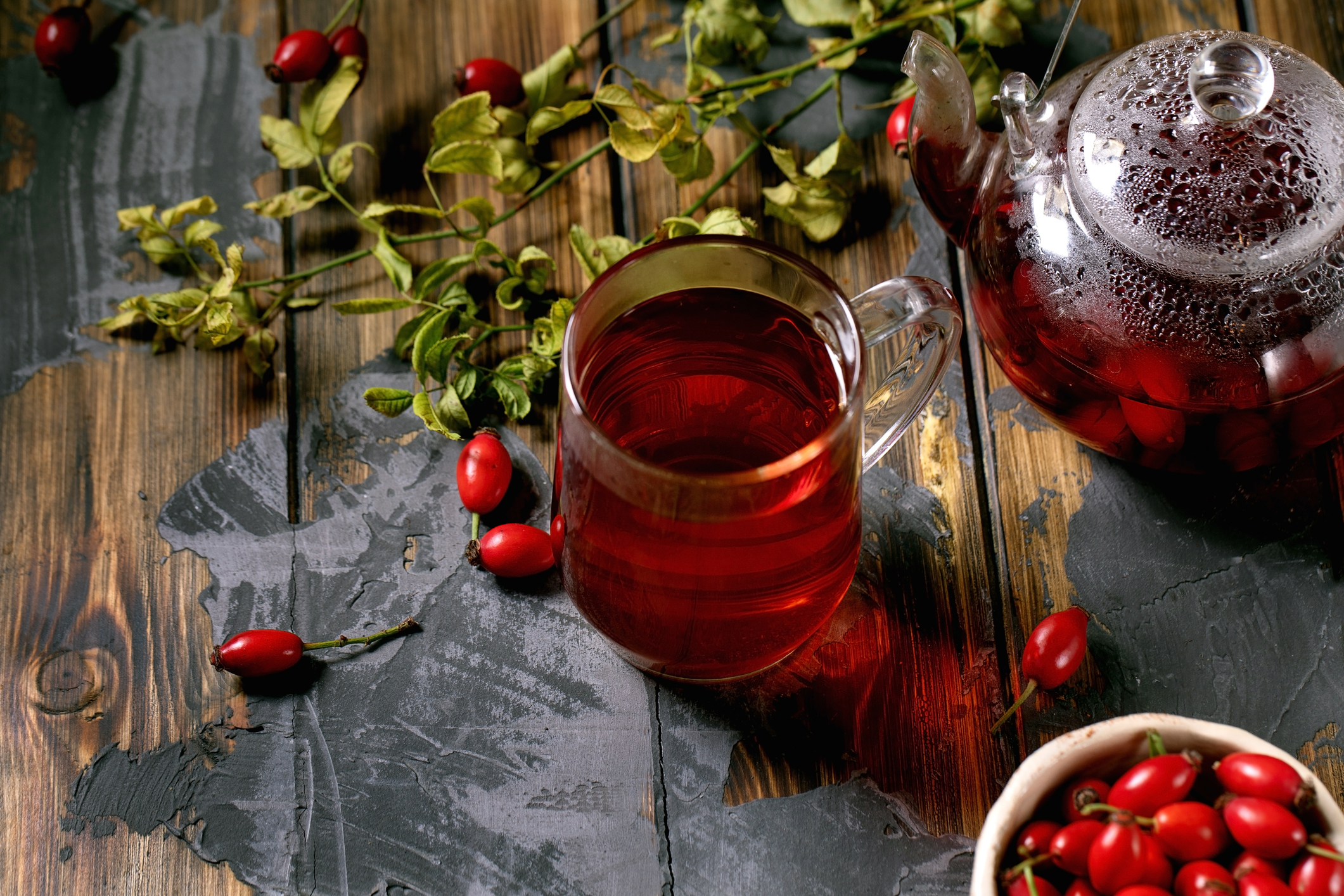 Cup of rose hip berries herbal tea and glass teapot standing on old wooden plank table with wild autumn berries around.