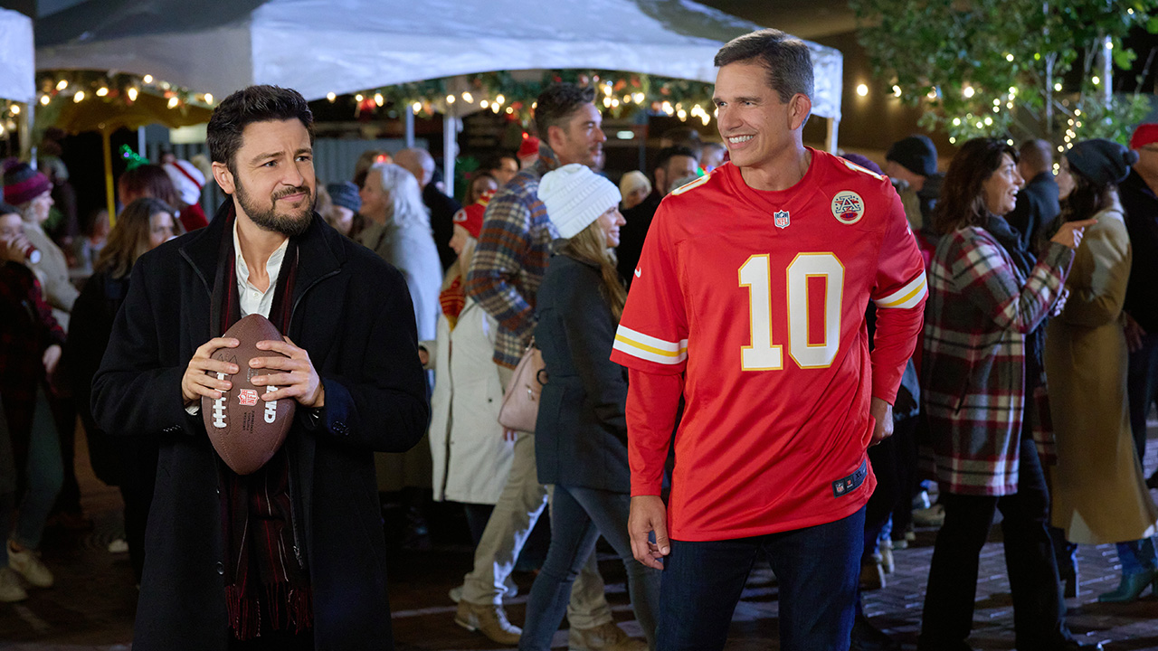 Trent Green, in a number 10 red Chiefs jersey, watches as a man in an overcoat with black short hair and a black beard holds a football, ready to throw it