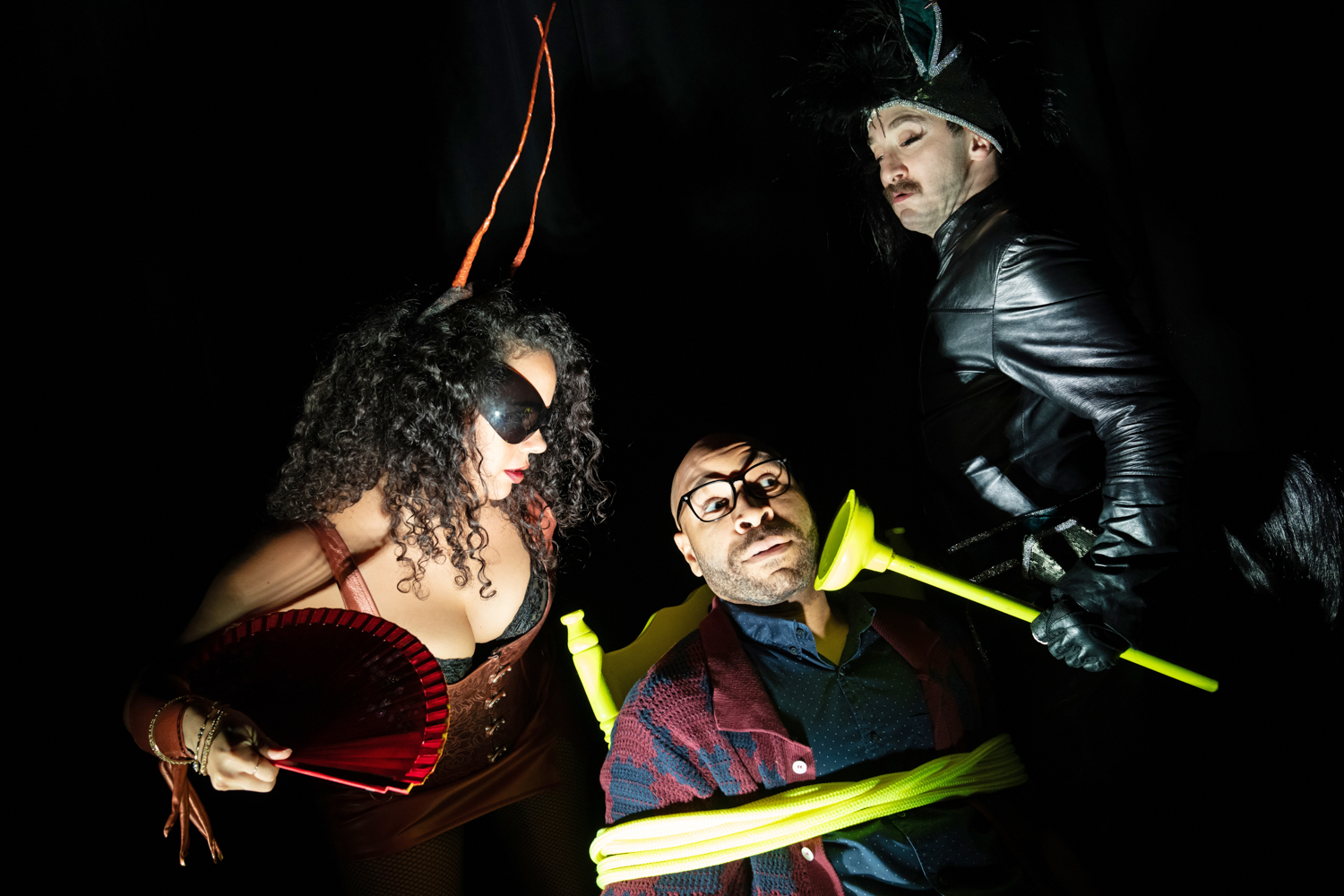 Still from Carmelita Tropicana. A person in sunglasses and a person in leather holding a neon plunger interrogate a man tied to a chair.