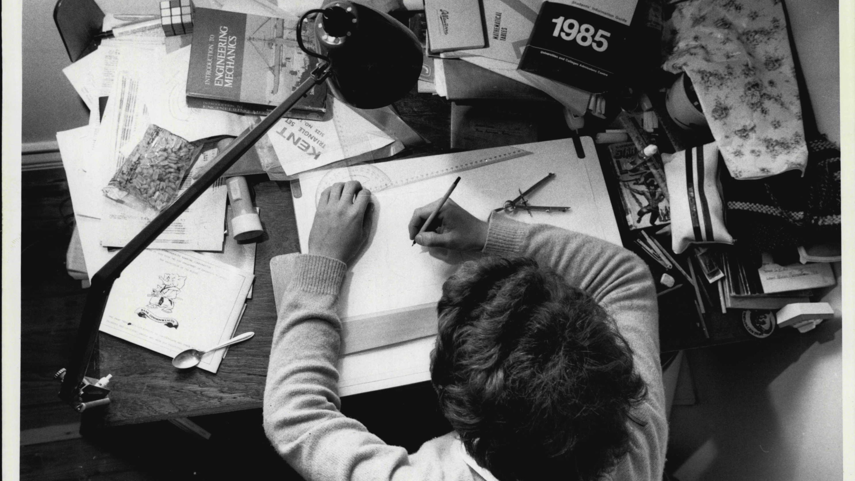 A person sits at a desk filled with books and writes in a notebook.