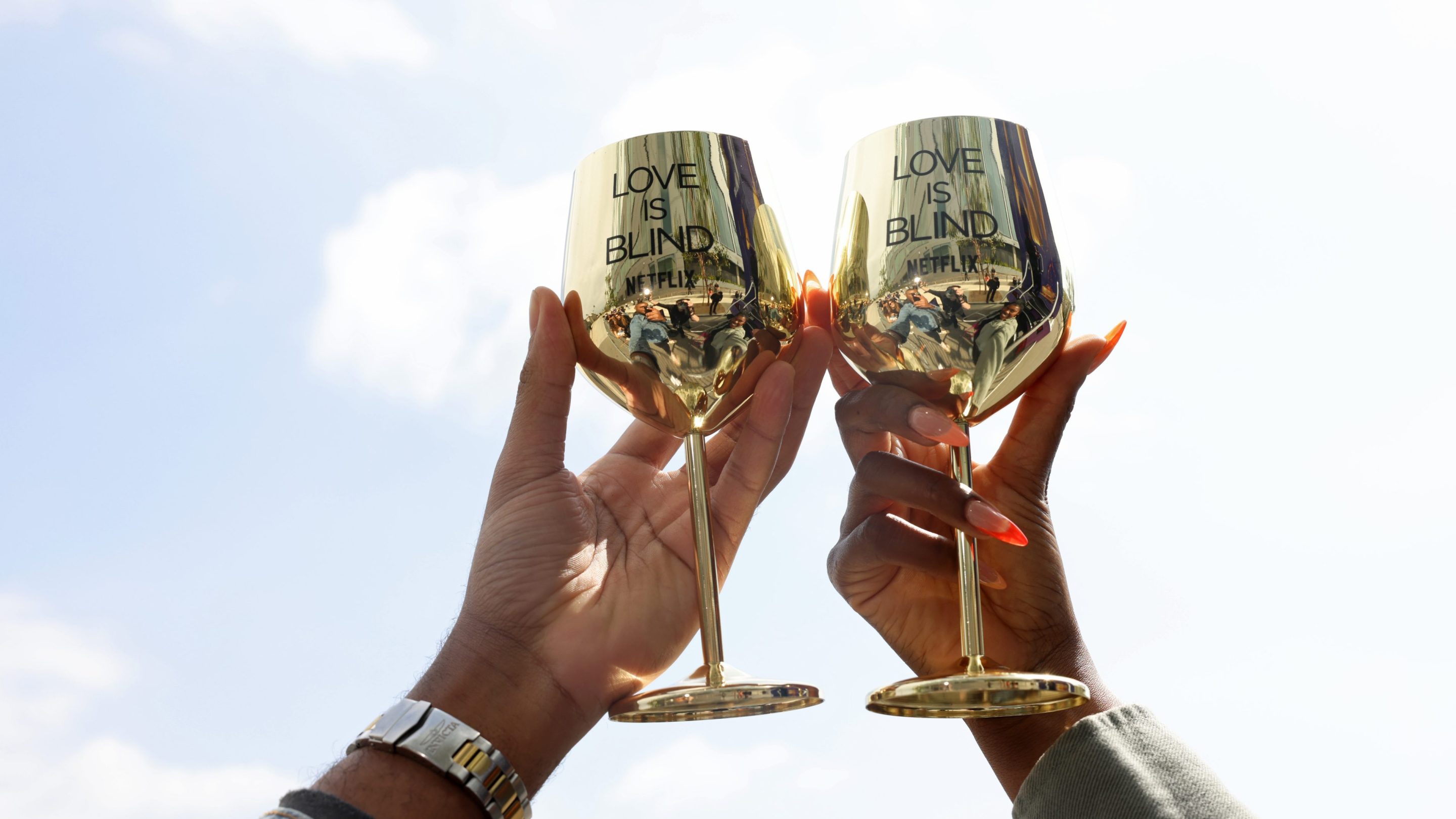 Two reflective gold wine glasses emblazoned with "Love is Blind" are held up against the sky.