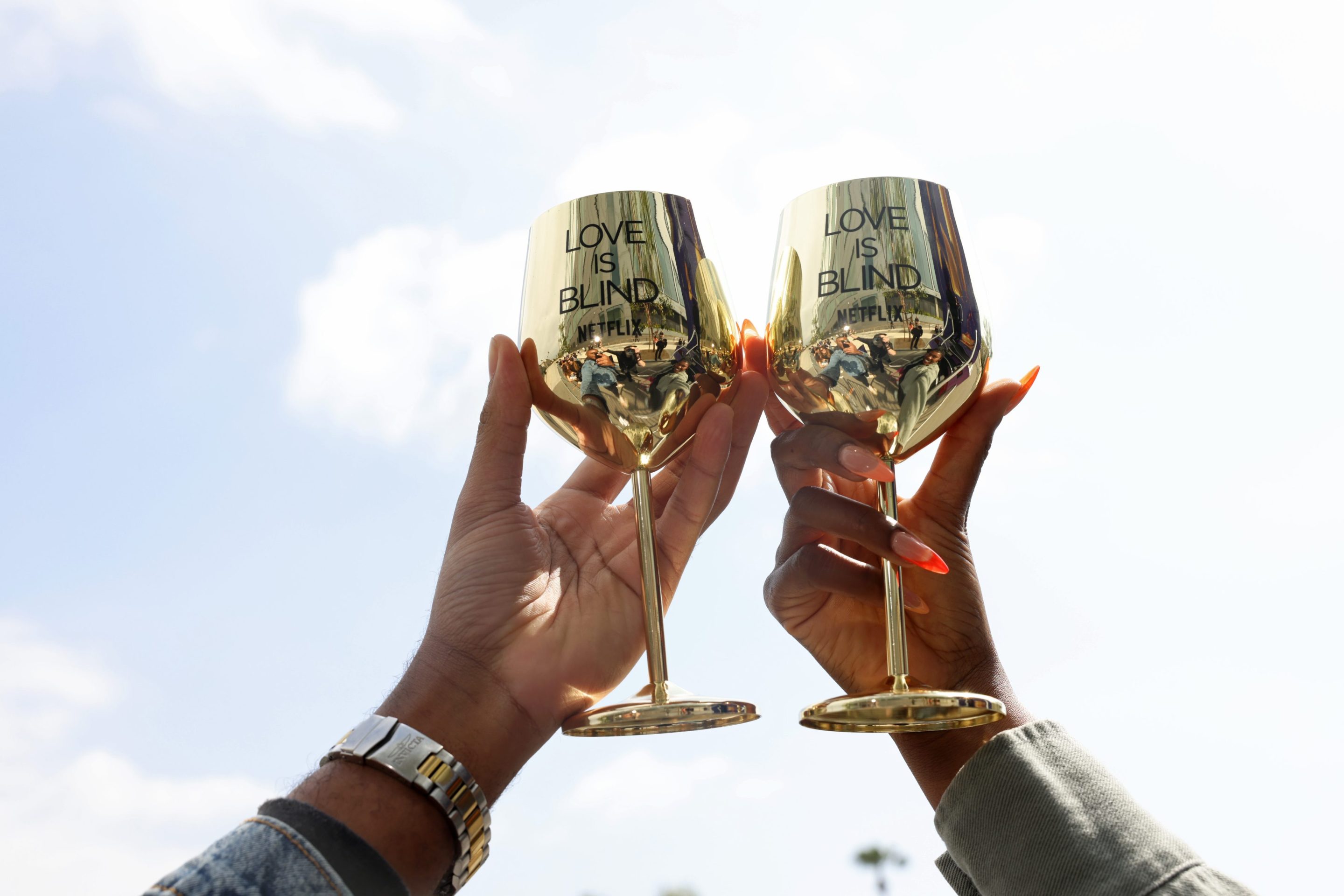 Two reflective gold wine glasses emblazoned with "Love is Blind" are held up against the sky.