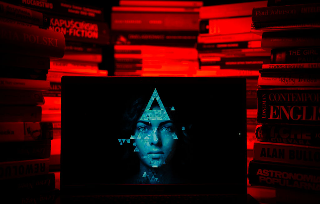 An artistic interpretation of artificial intelligence, showing a stack of books surrounding a laptop with a pixelated face on it