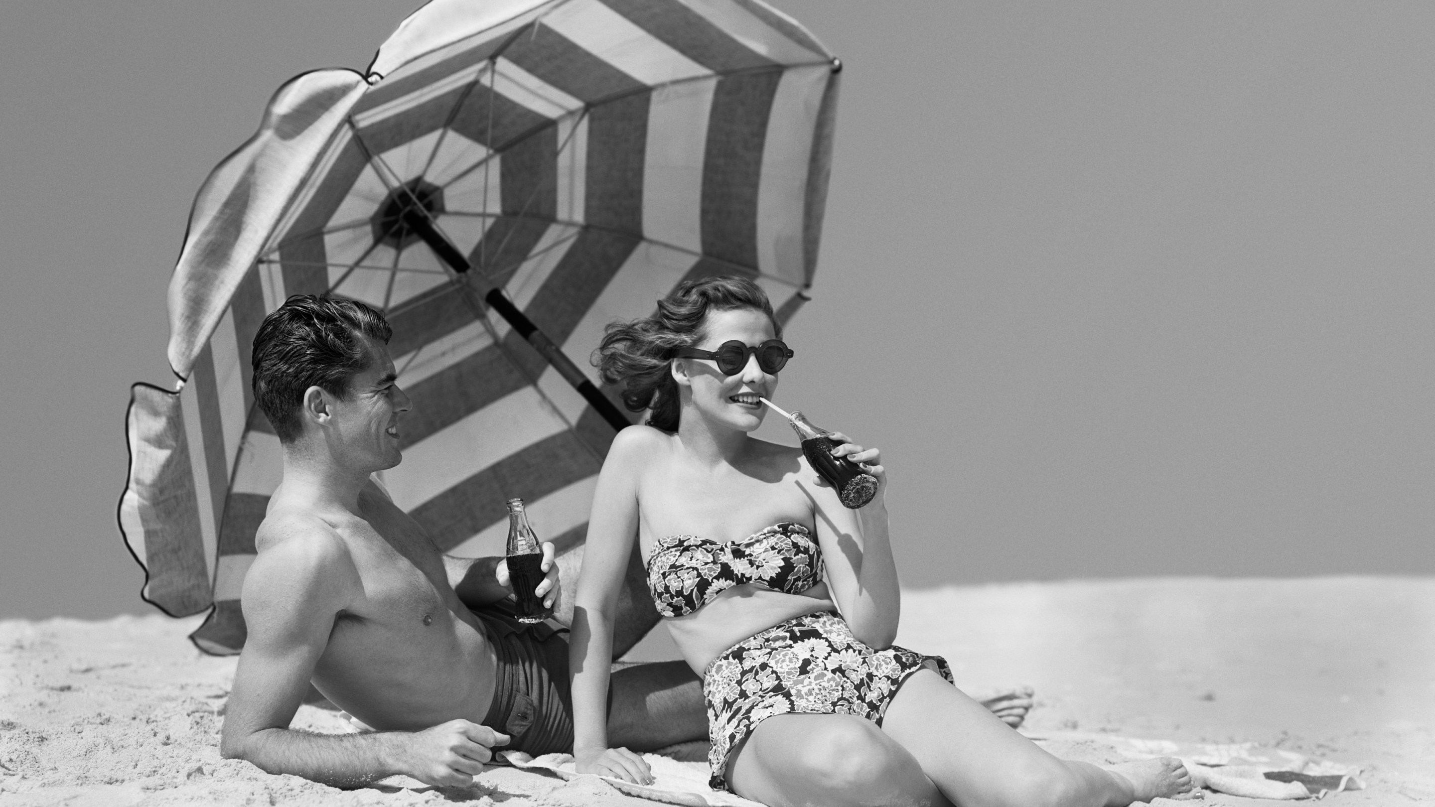 A man and woman sit on a beach under a striped umbrella drinking Coke out of glass bottles.