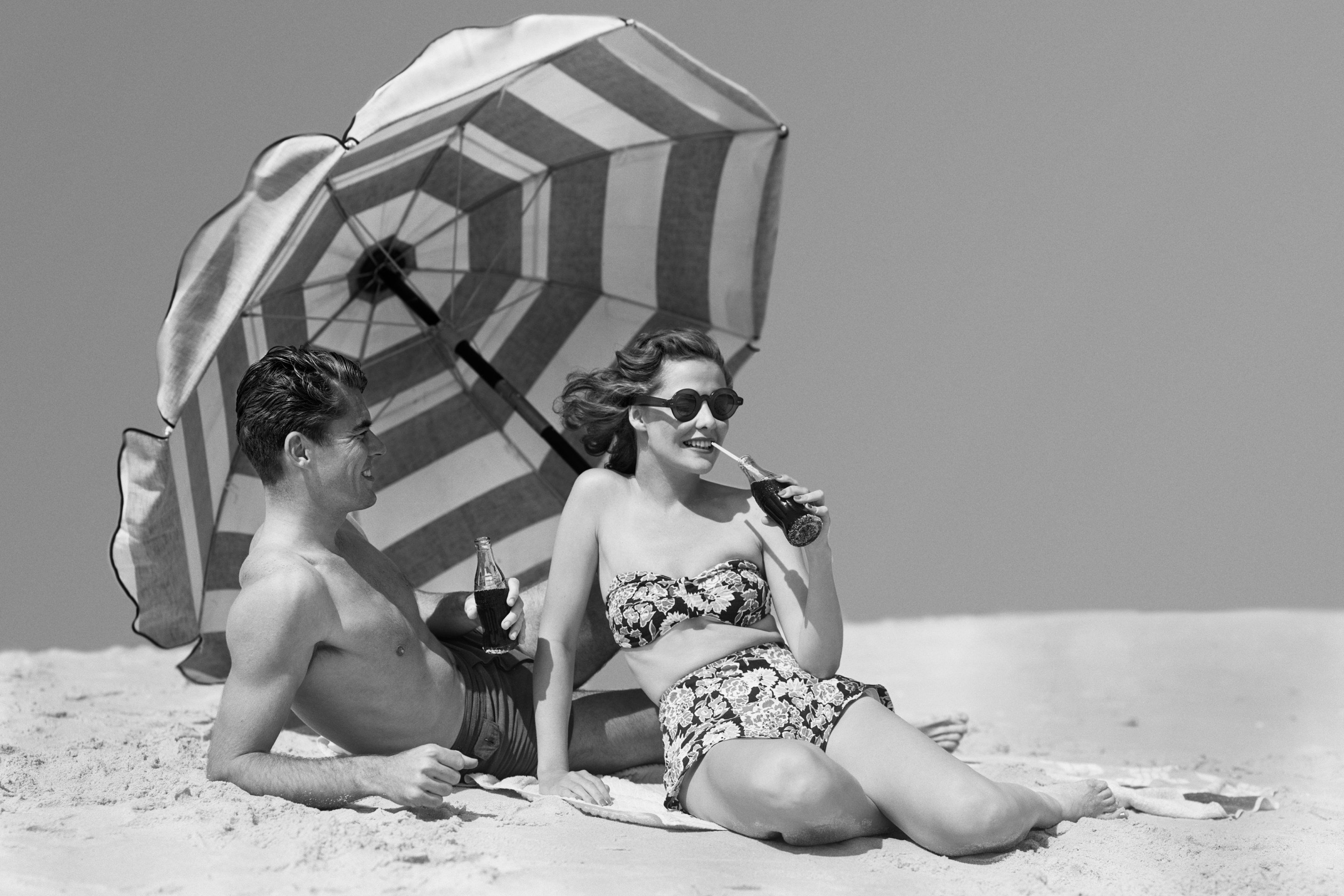 A man and woman sit on a beach under a striped umbrella drinking Coke out of glass bottles.