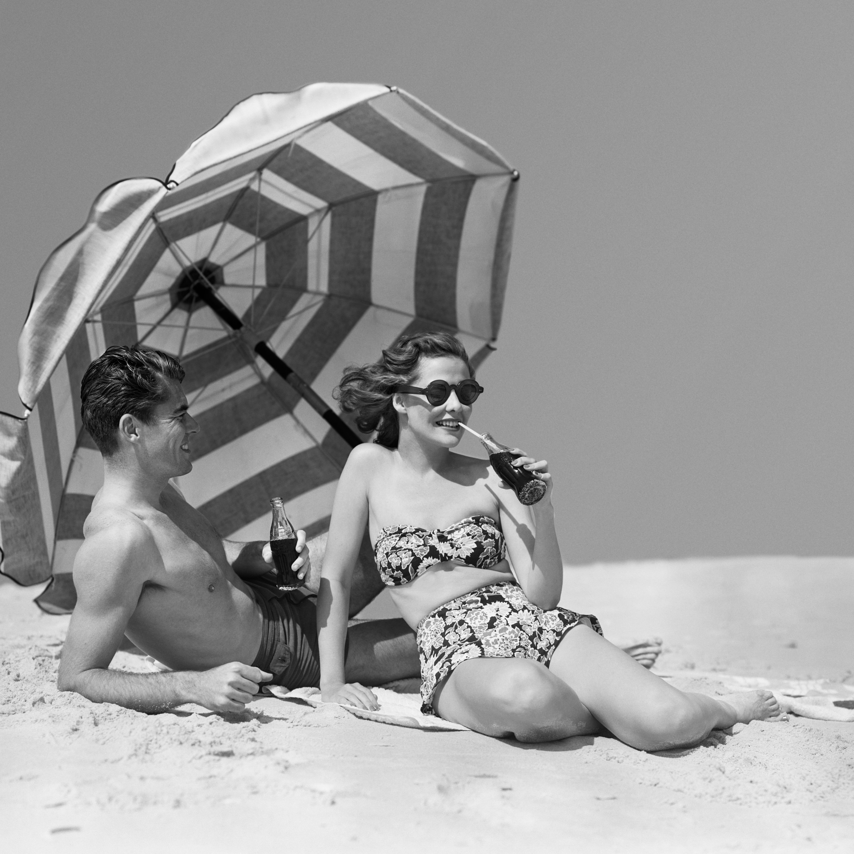 A man and woman sit on a beach under a striped umbrella drinking Coke out of glass bottles.