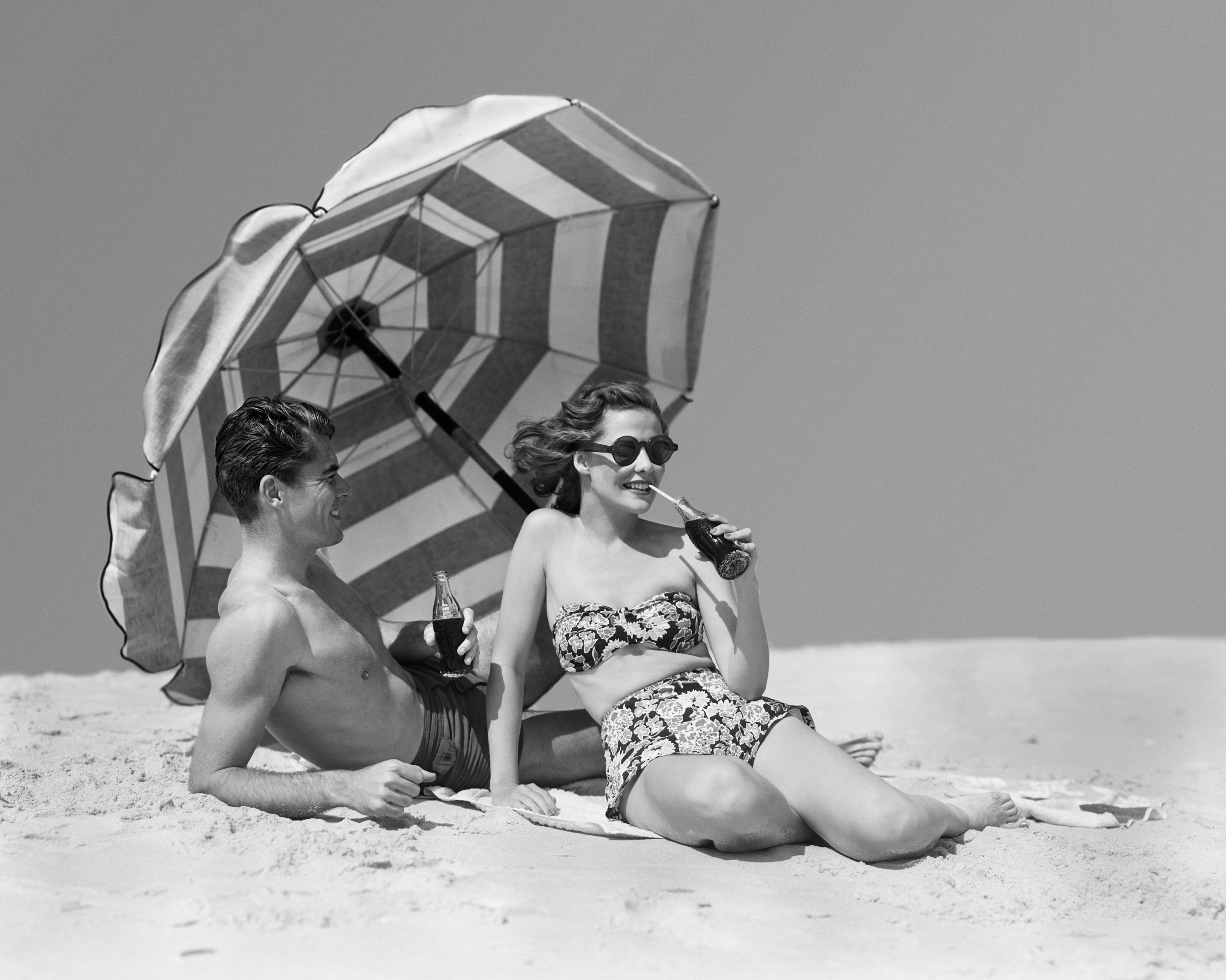 A man and woman sit on a beach under a striped umbrella drinking Coke out of glass bottles.