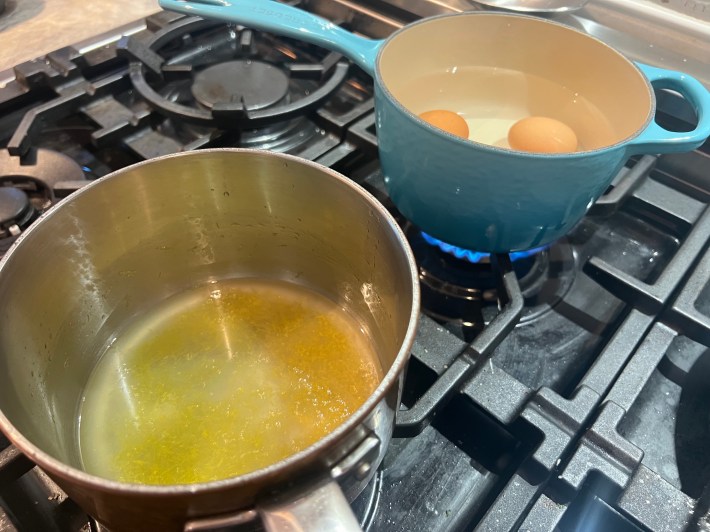 In the foreground, lemon juice, lemon zest, and sugar simmer in a saucepan. In the background, three eggs are submerged in water in another saucepan.