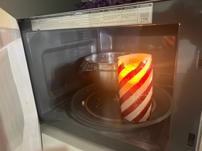 A striped Christmas pillar candle, lit, and a metal mixing bowl sit inside a small microwave.