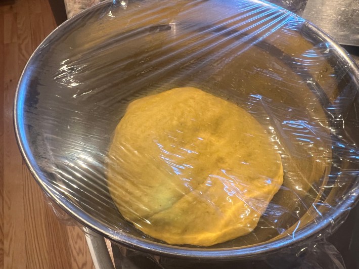 A blob of bread dough under plastic film in a large metal mixing bowl.