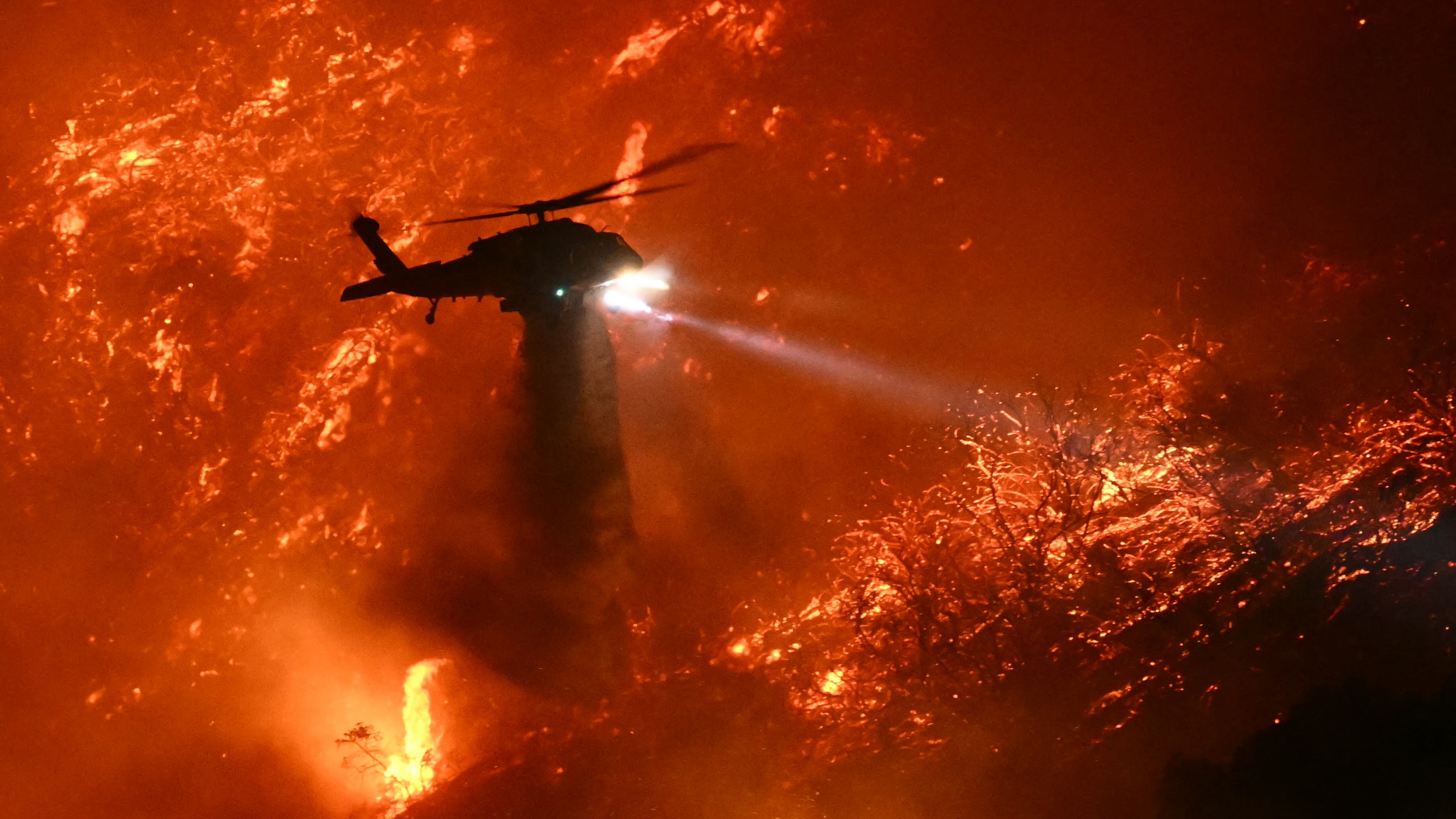 A fire fighting helicopter drops water as the Palisades fire grows near the Mandeville Canyon neighborhood and Encino, California, on January 11, 2025. The Palisades Fire, the largest of the Los Angeles fires, spread toward previously untouched neighborhoods January 11, forcing new evacuations and dimming hopes that the disaster was coming under control. Across the city, at least 11 people have died as multiple fires have ripped through residential areas since January 7, razing thousands of homes in destruction that US President Joe Biden likened to a "war scene." (Photo by Patrick T. Fallon / AFP) (Photo by PATRICK T. FALLON/AFP via Getty Images)