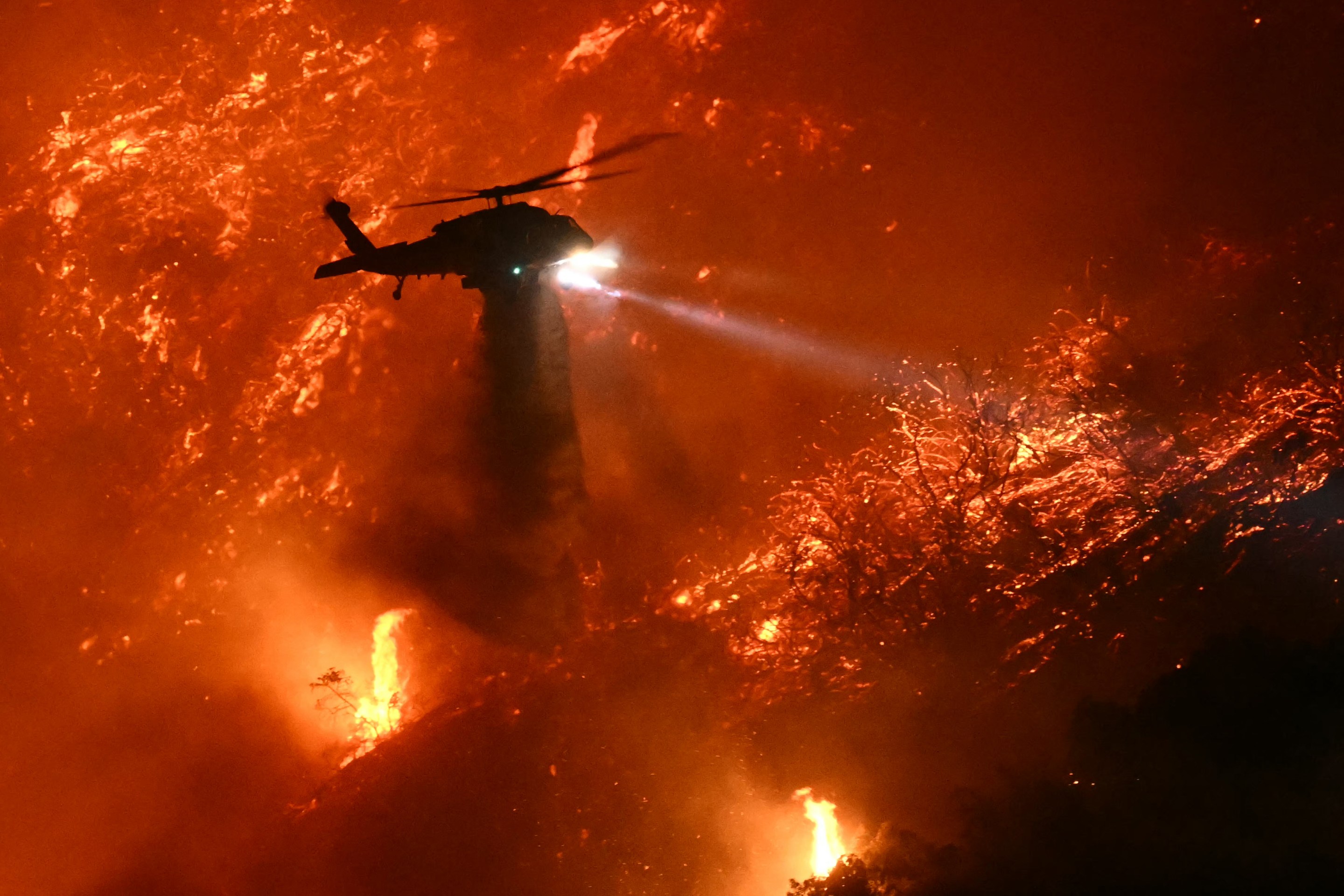 A fire fighting helicopter drops water as the Palisades fire grows near the Mandeville Canyon neighborhood and Encino, California, on January 11, 2025. The Palisades Fire, the largest of the Los Angeles fires, spread toward previously untouched neighborhoods January 11, forcing new evacuations and dimming hopes that the disaster was coming under control. Across the city, at least 11 people have died as multiple fires have ripped through residential areas since January 7, razing thousands of homes in destruction that US President Joe Biden likened to a "war scene." (Photo by Patrick T. Fallon / AFP) (Photo by PATRICK T. FALLON/AFP via Getty Images)