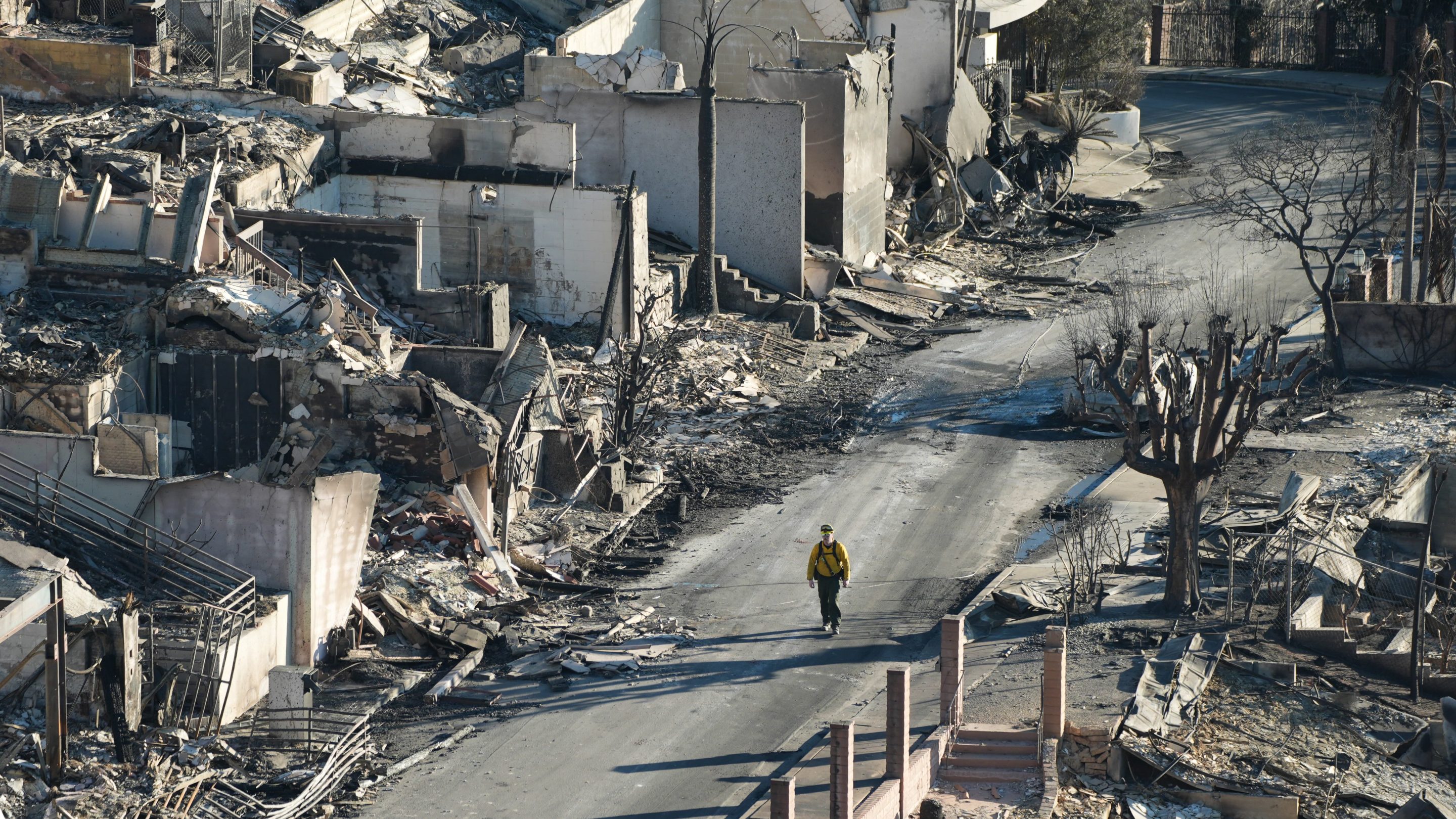 An aerial view of a street in the Eaton fire. The entire street is burned and decimated. It looks like a bomb went off. A sole person walks down the empty steet.