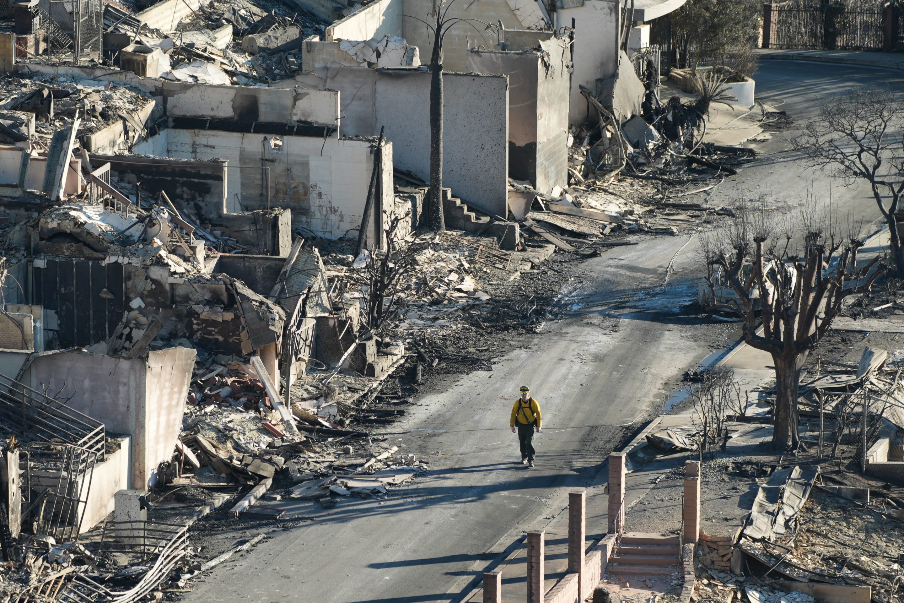 An aerial view of a street in the Eaton fire. The entire street is burned and decimated. It looks like a bomb went off. A sole person walks down the empty steet.