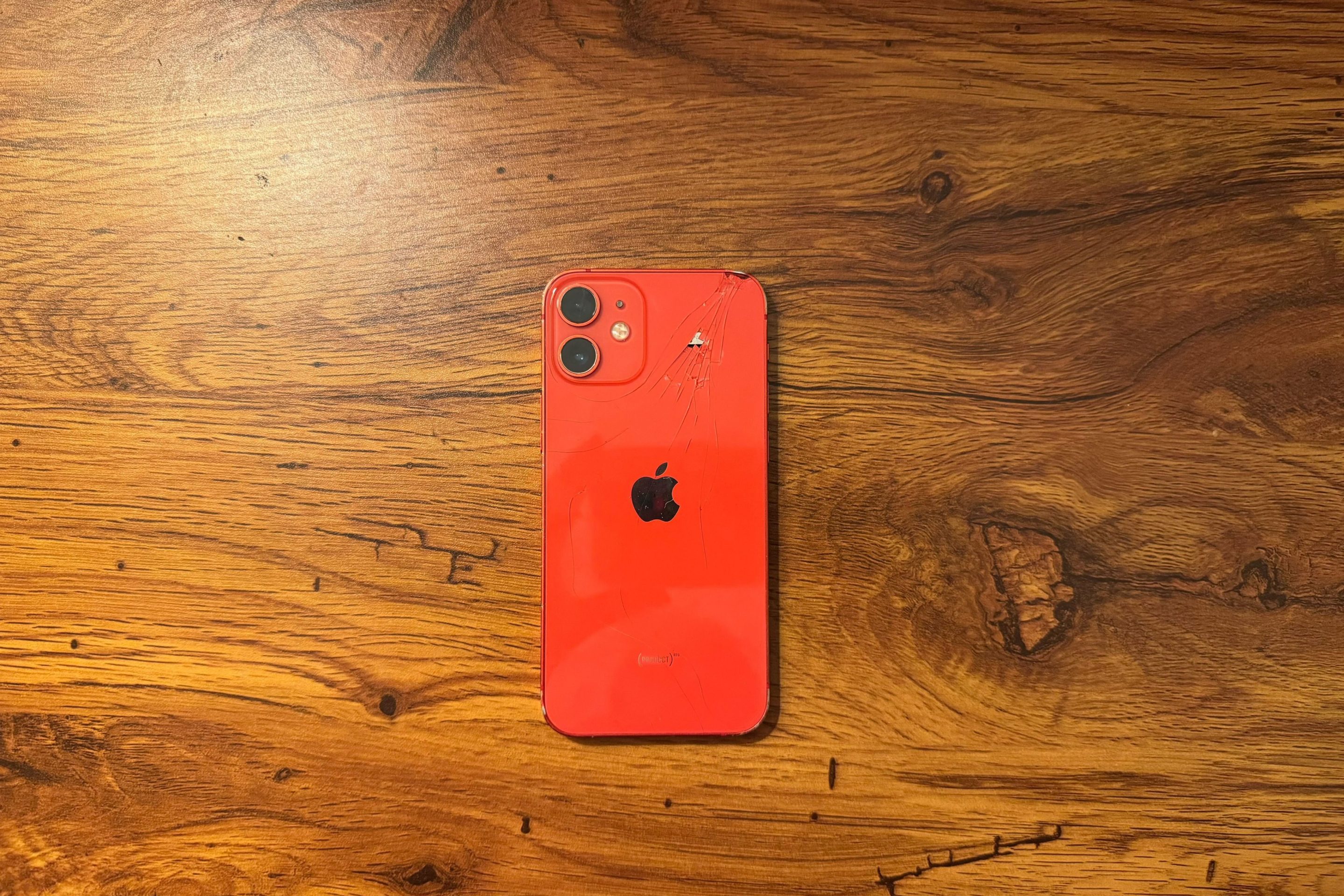 A red iPhone 12 mini rests on a wood table. The back of the phone is facing up; it's glass surface is cracked.